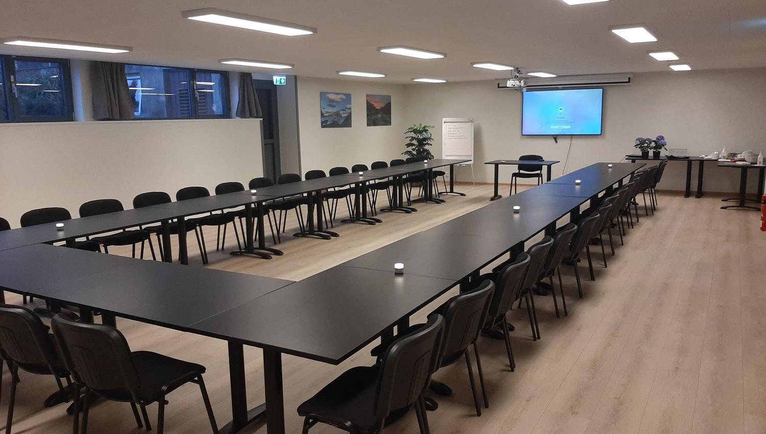 Stort møterom med U-forma svarte bord og stolar, skjerm på veggen, flippover og serveringsbord langs sida..