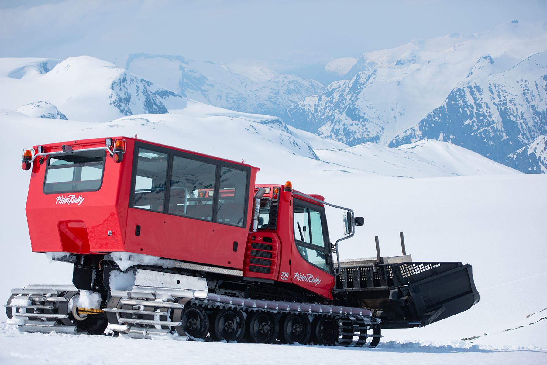 A red tracked snowcat with a front plow sits on a snowy slope, with distant snow-covered mountains behind it.