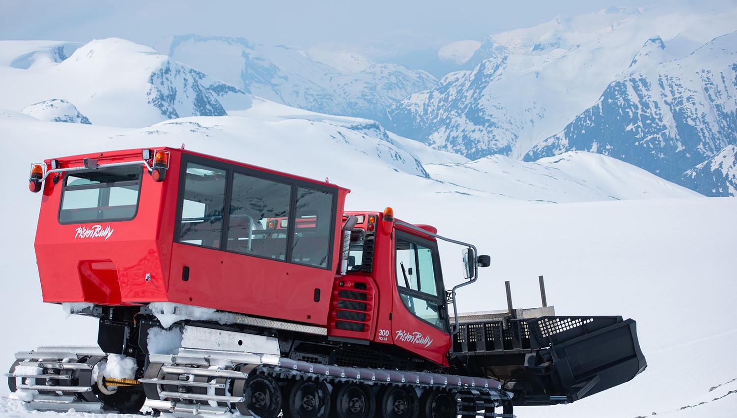 A red tracked snowcat with a front plow sits on a snowy slope, with distant snow-covered mountains behind it.