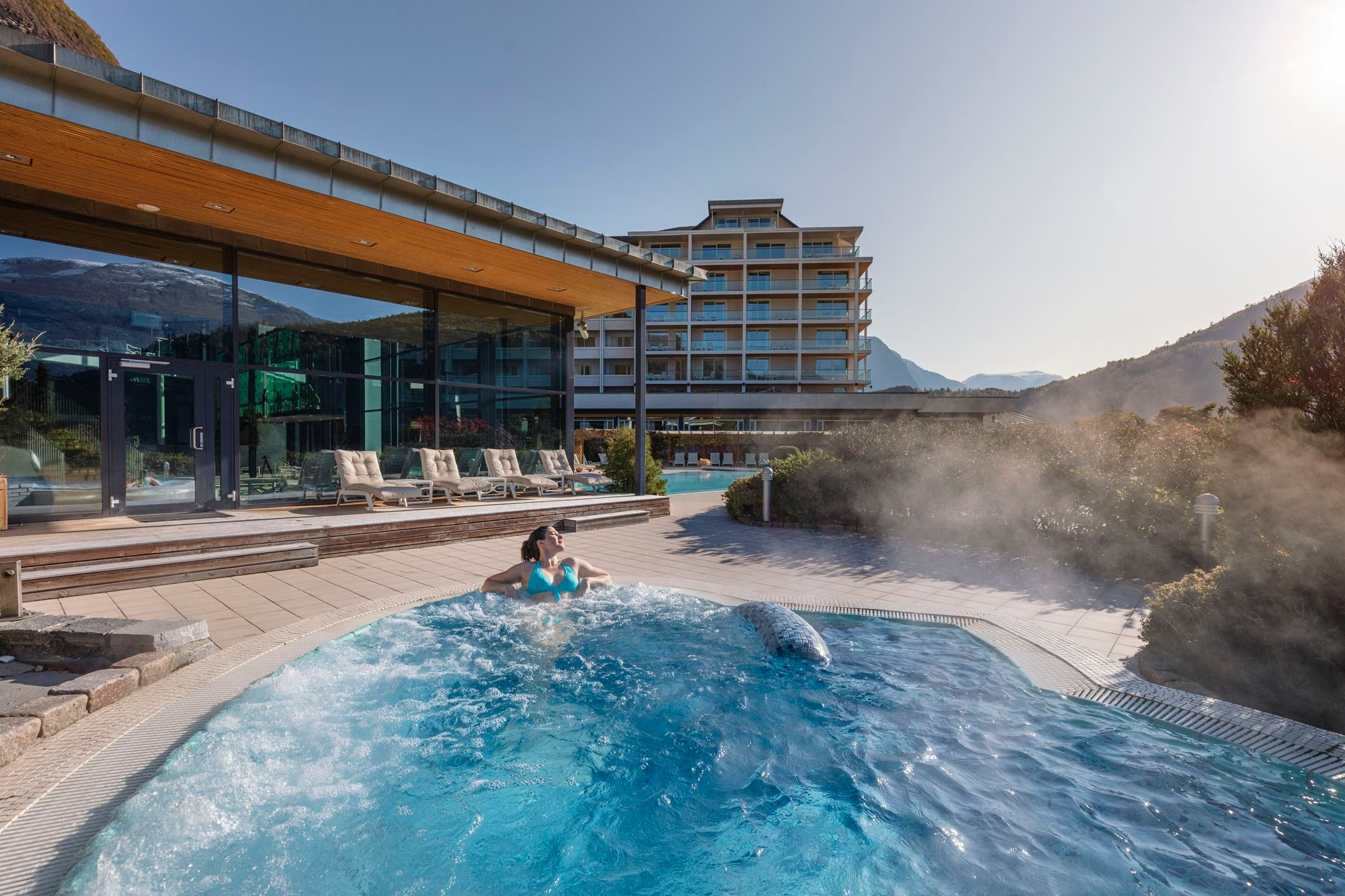 Person relaxing in a bubbling outdoor hot tub beside a modern glass building, with mountains and steam in sunlight..