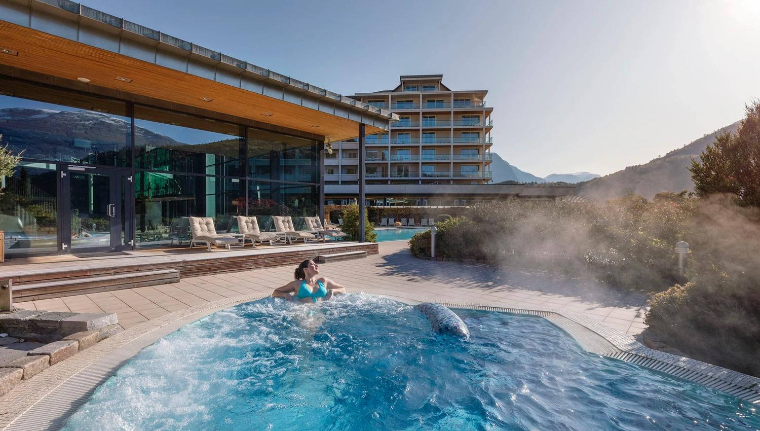 Person relaxing in a bubbling outdoor hot tub beside a modern glass building, with mountains and steam in sunlight..