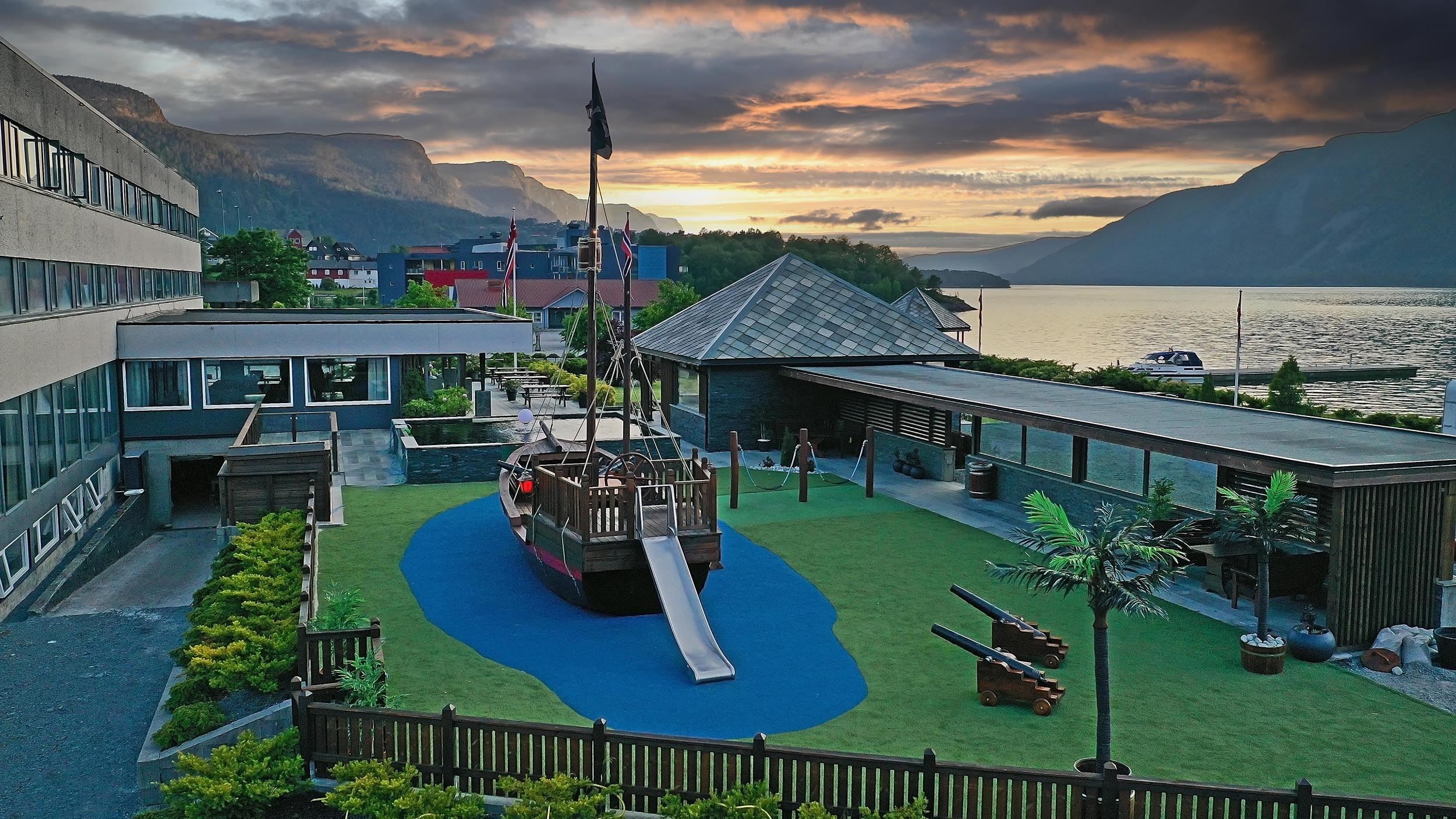 Uteområde ved fjord med sjørøvarskute-leikeapparat, sklie, kunstgras, bygningar og fjell i solnedgang..