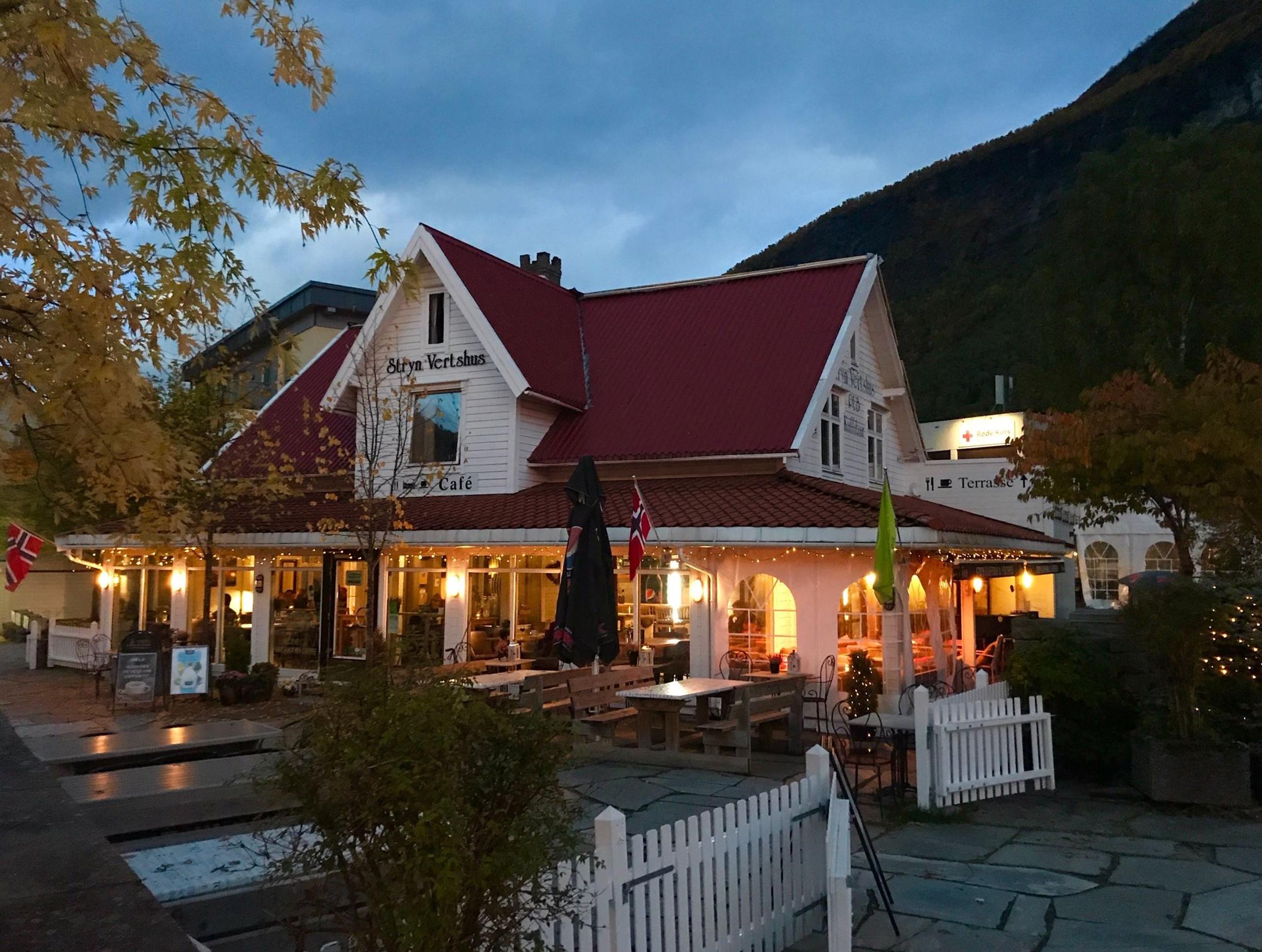 A warmly lit café with a red roof and outdoor seating, framed by autumn trees and mountains under a cloudy sky.