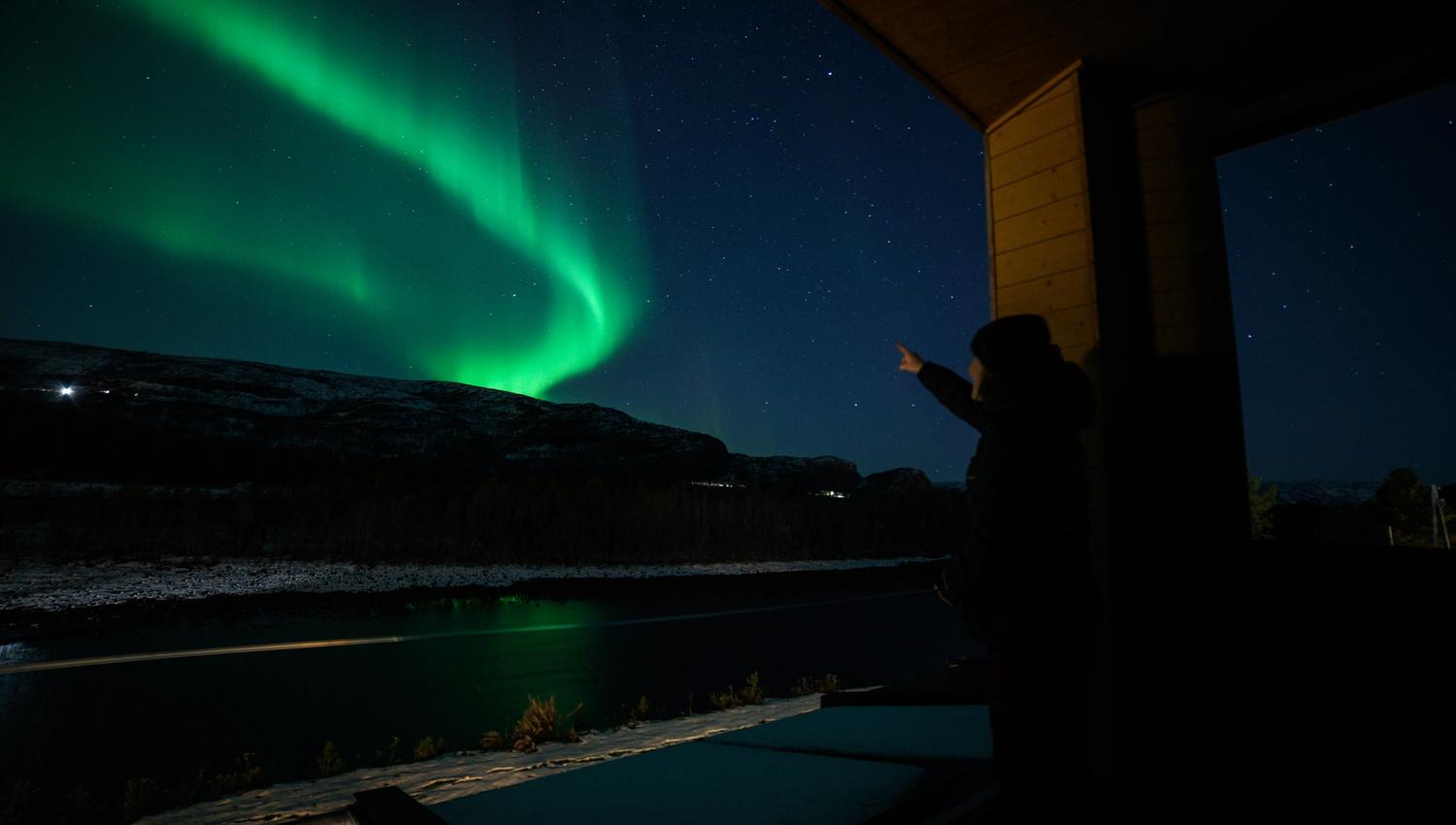 Experience the magic of the Northern Lights! This breathtaking view captures a person pointing to the vibrant green auroras illuminating the night sky, set against a serene snowy landscape and twinkling stars. Discover the beauty of nature's light show!