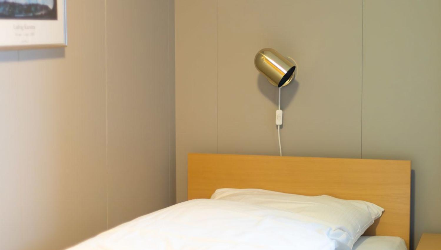 A neatly made single bed with white bedding sits below a small wall lamp, beside a nightstand and framed art..