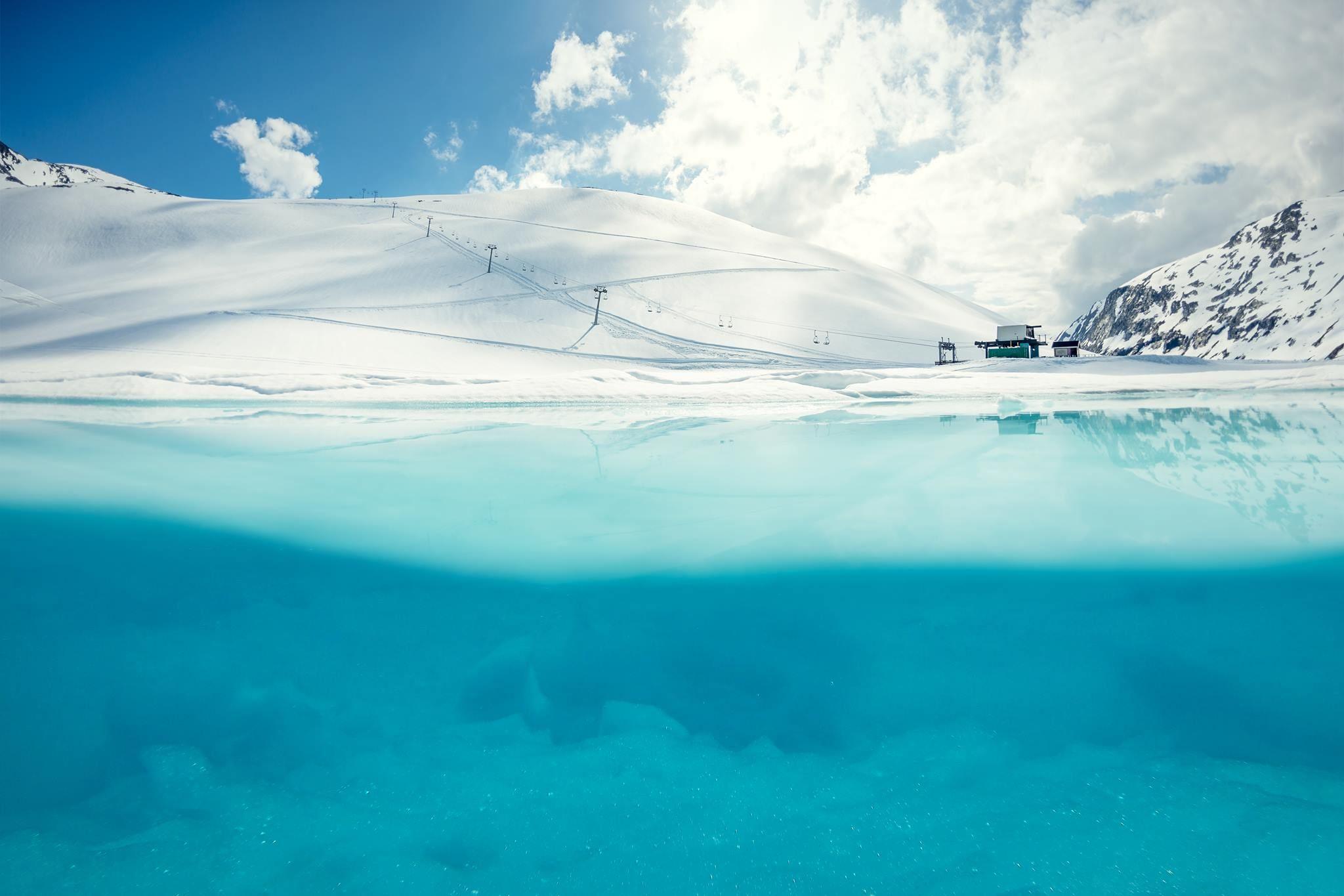 Snow-covered mountain with ski lift towers and a small building, above a clear turquoise lake under clouds.
