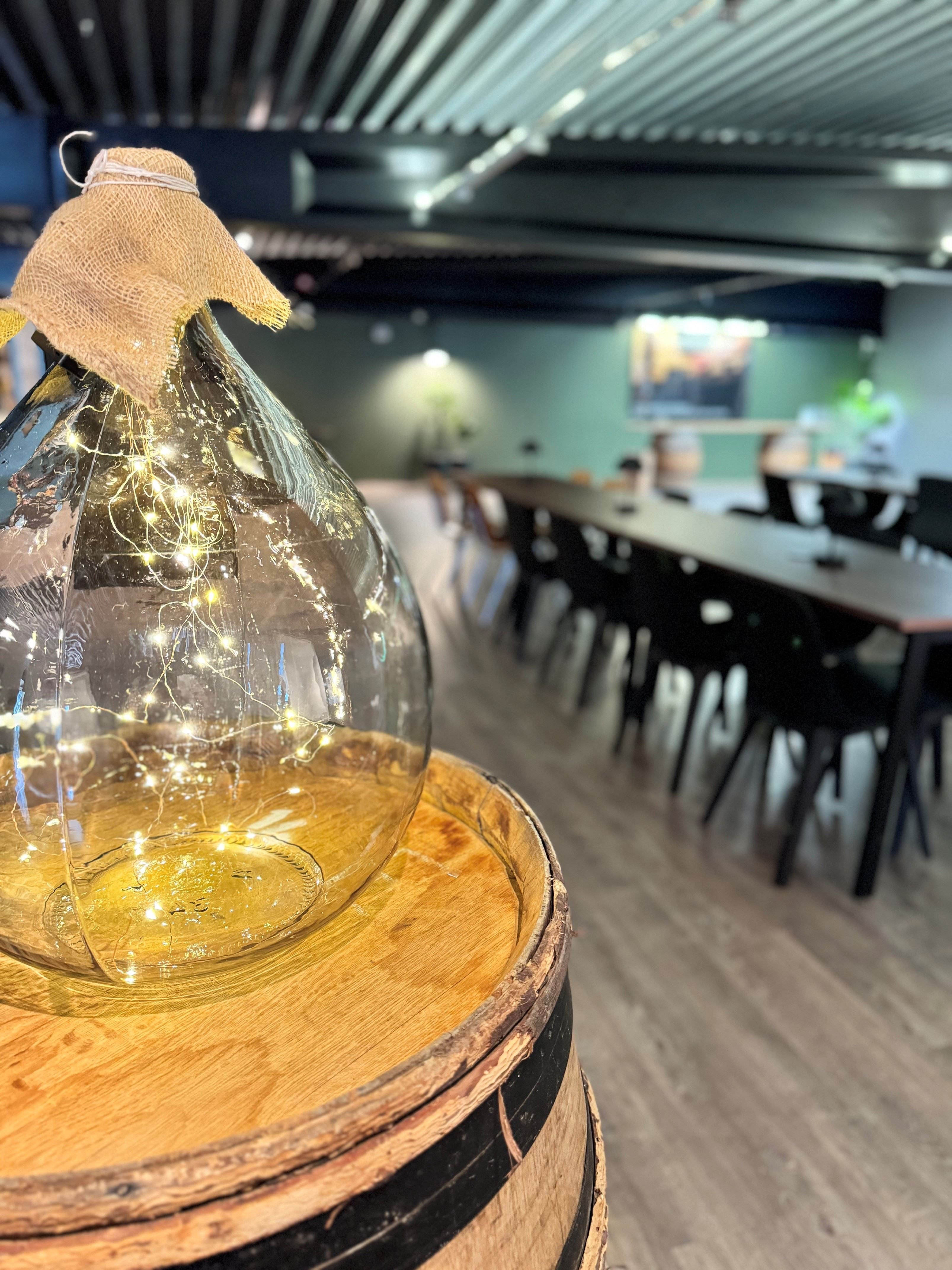 Large clear glass bottle filled with string lights on a wooden barrel, with a blurred dining area in the background.