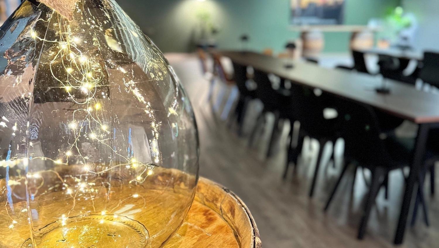 Large clear glass bottle filled with string lights on a wooden barrel, with a blurred dining area in the background.