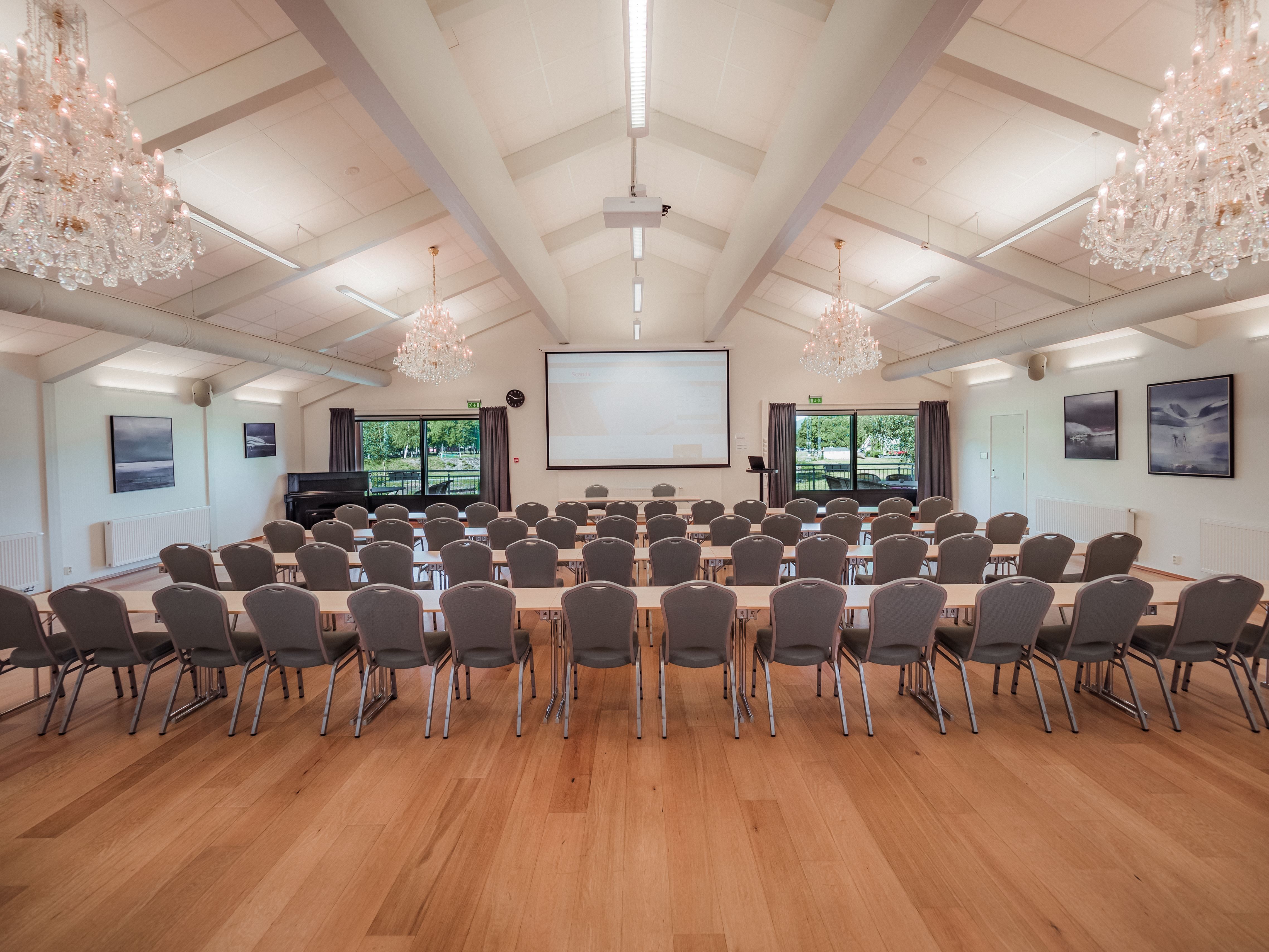 Empty meeting hall with rows of chairs and tables facing a projection screen, under chandeliers and ceiling lights.