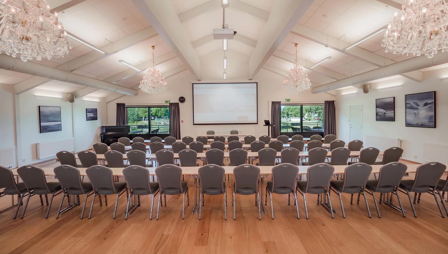 Empty meeting hall with rows of chairs and tables facing a projection screen, under chandeliers and ceiling lights.