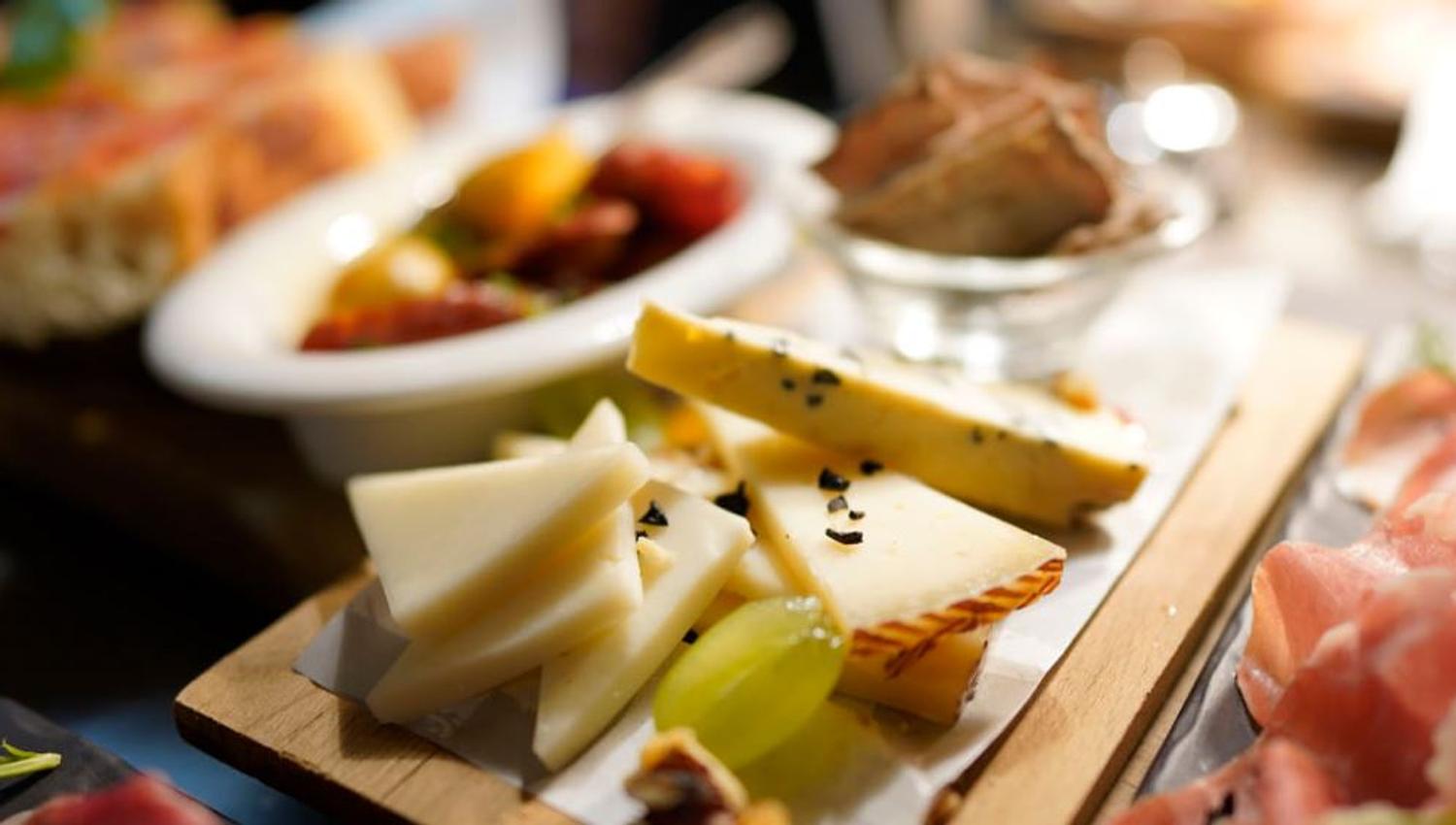 Nærbilde av ost med sorte frø, grønne druer og kjeks, med spekemat og bruschetta i bakgrunnen.