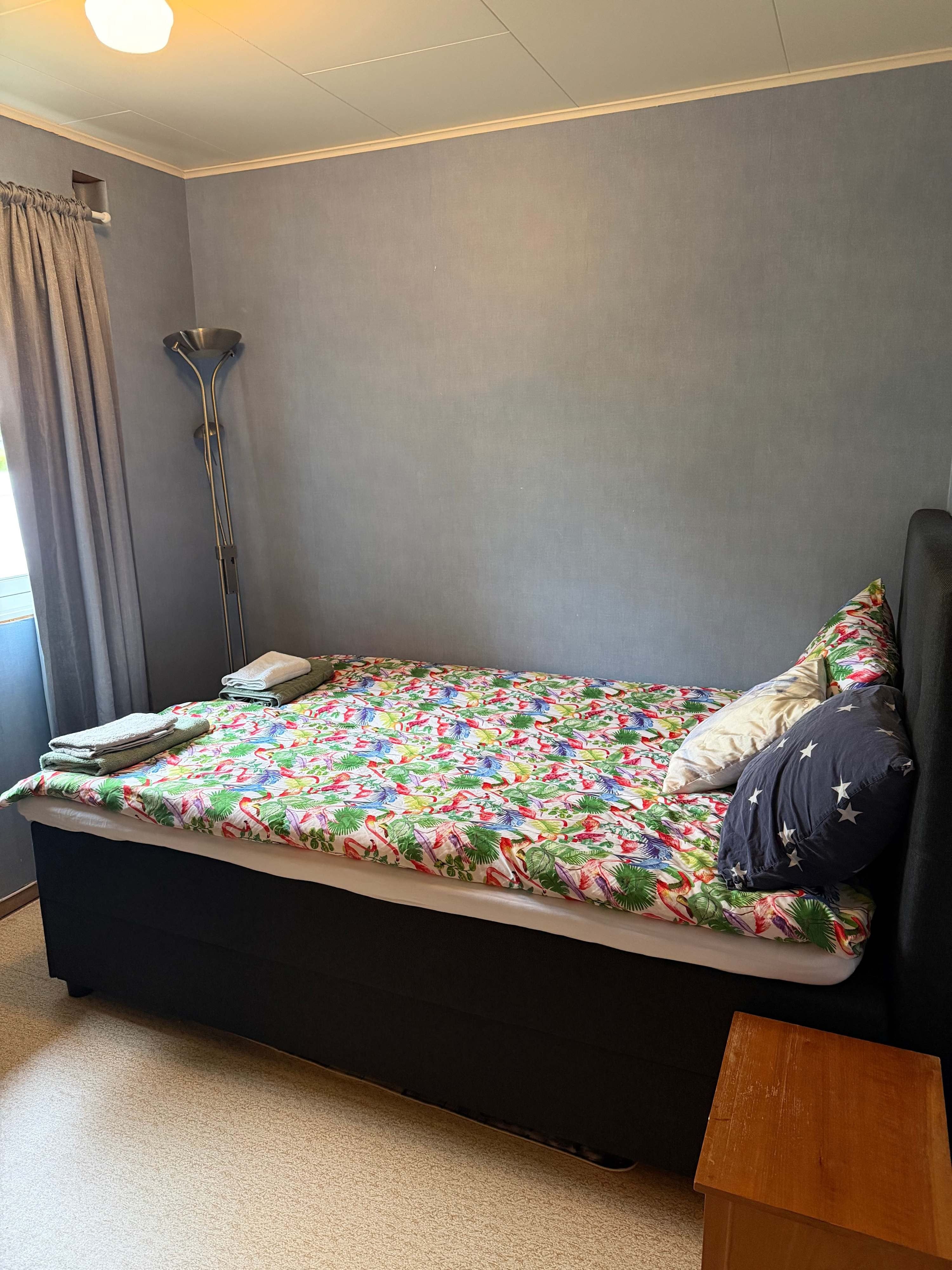 Cozy bedroom with a double bed, floral bedspread, stacked towels, floor lamp, and gray curtains by a window.