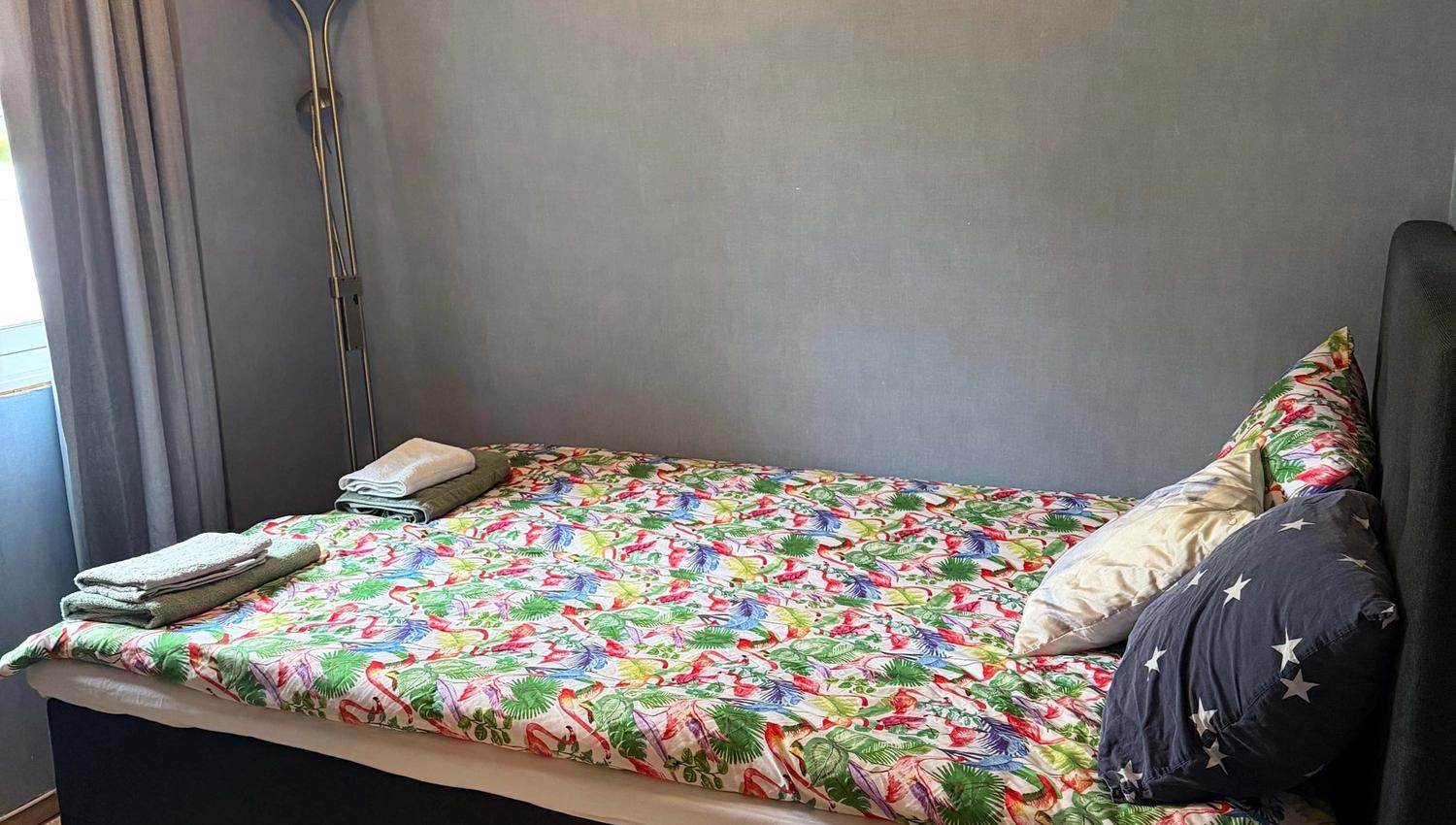 Cozy bedroom with a double bed, floral bedspread, stacked towels, floor lamp, and gray curtains by a window.