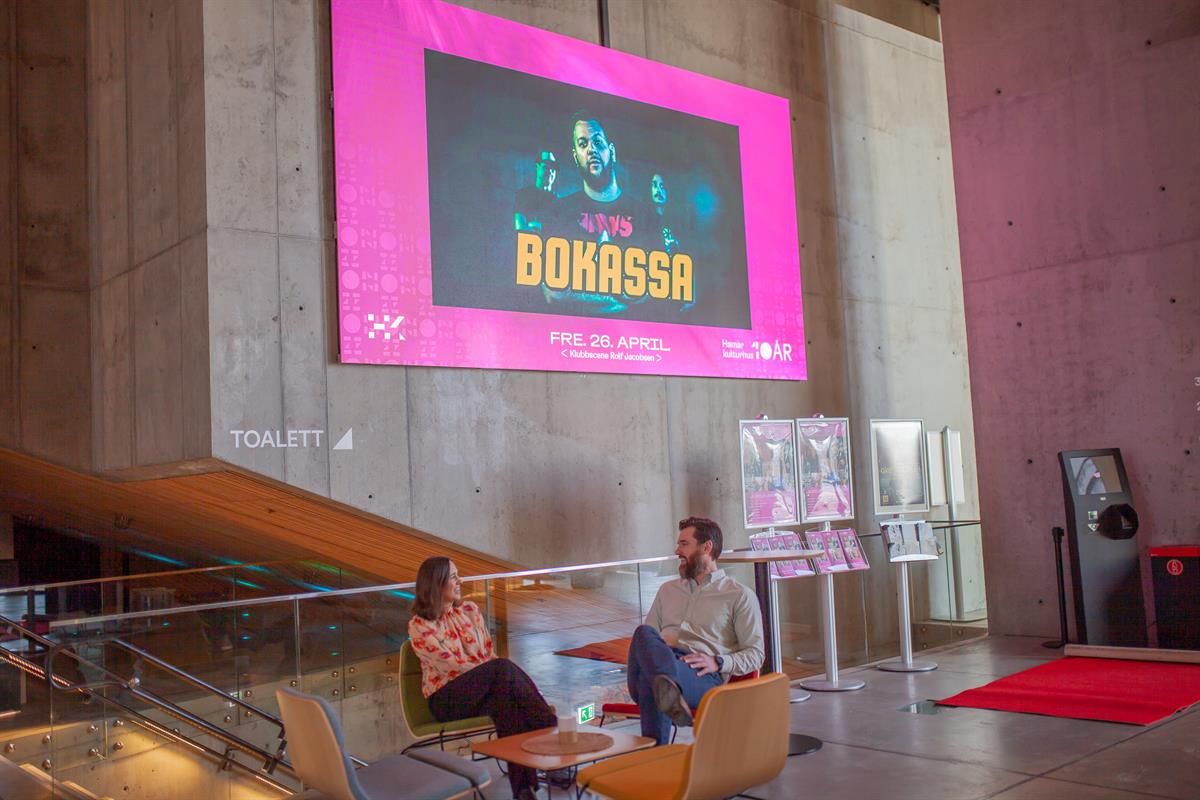 To personer sitter i en åpen lounge- eller foajéområde i Hamar kulturhus, omgitt av moderne betongvegger og et stort, fargerikt skjermbanner som viser en kulturplakat.