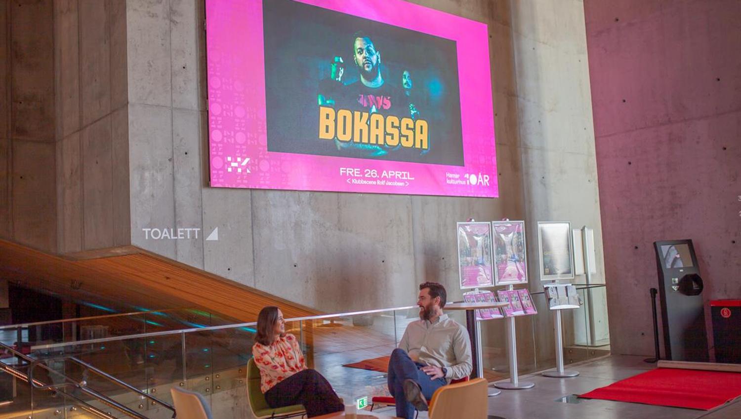 To personer sitter i en åpen lounge- eller foajéområde i Hamar kulturhus, omgitt av moderne betongvegger og et stort, fargerikt skjermbanner som viser en kulturplakat.