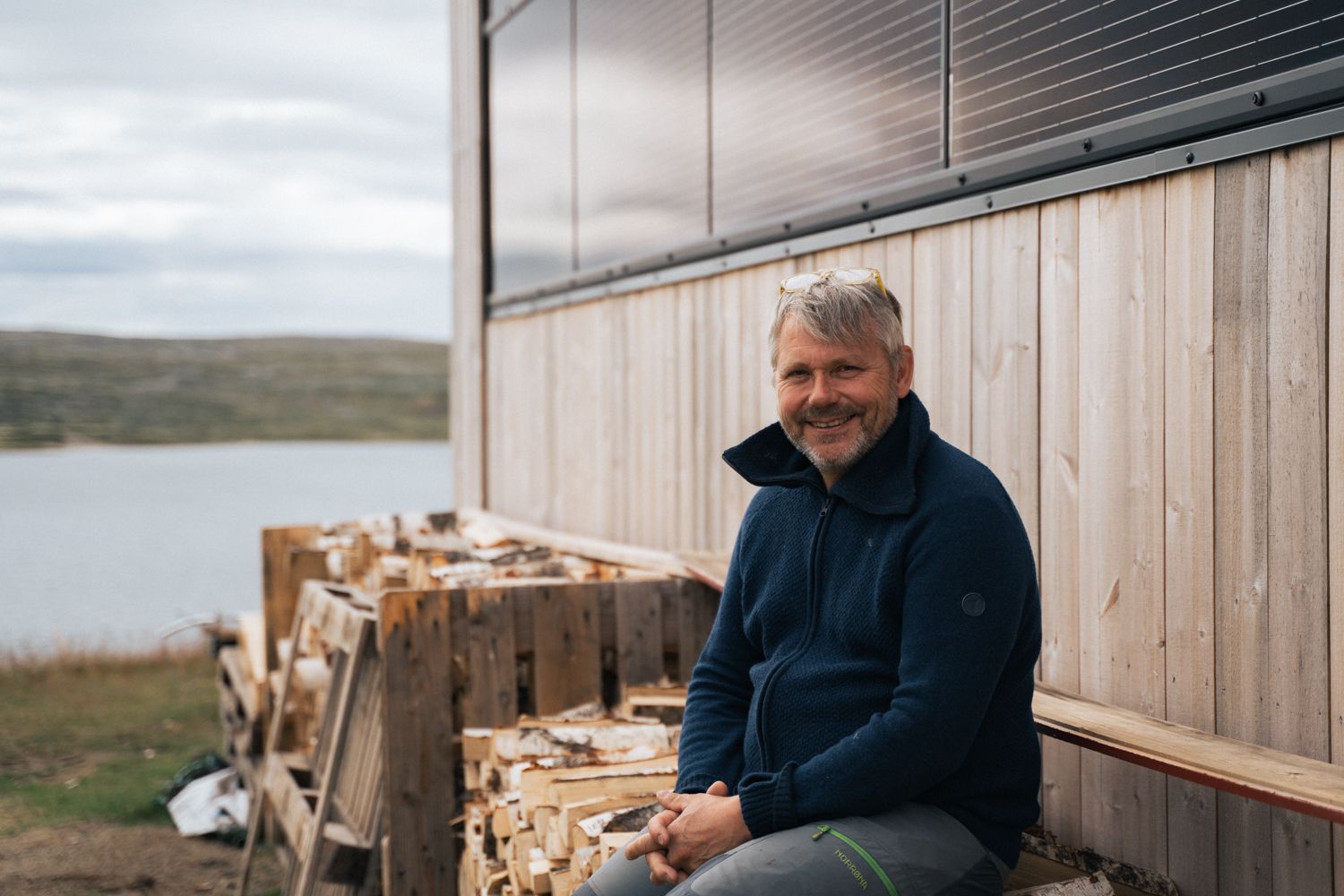 The manager at Rauhelleren sits on a woodpile and smiles at the photographer.