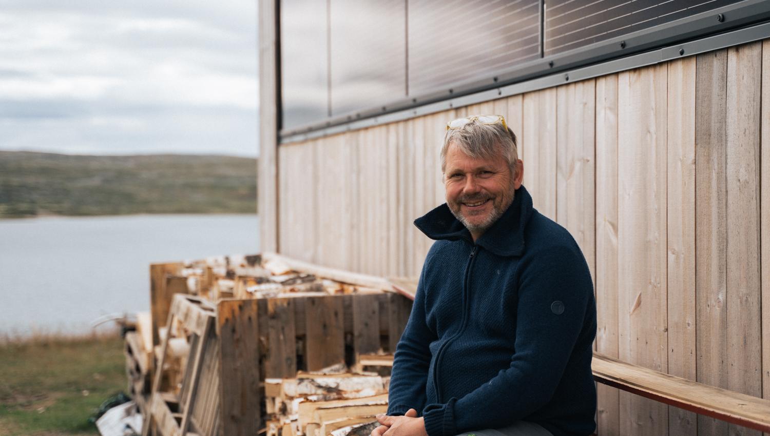 The manager at Rauhelleren sits on a woodpile and smiles at the photographer.