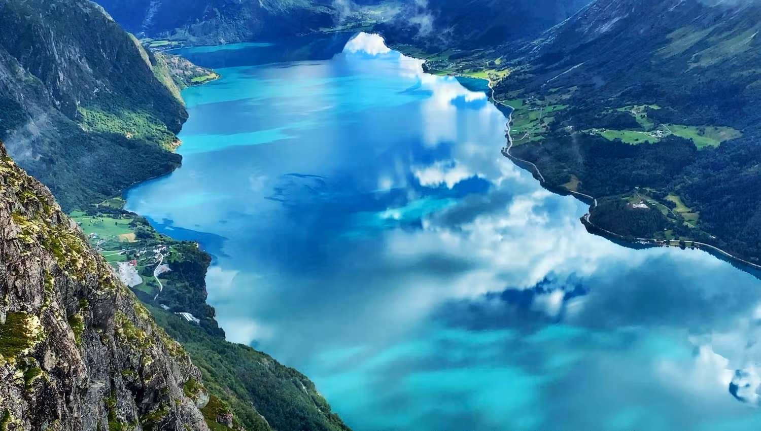 High viewpoint over a turquoise fjord surrounded by steep green mountains, rocky foreground, and low clouds..