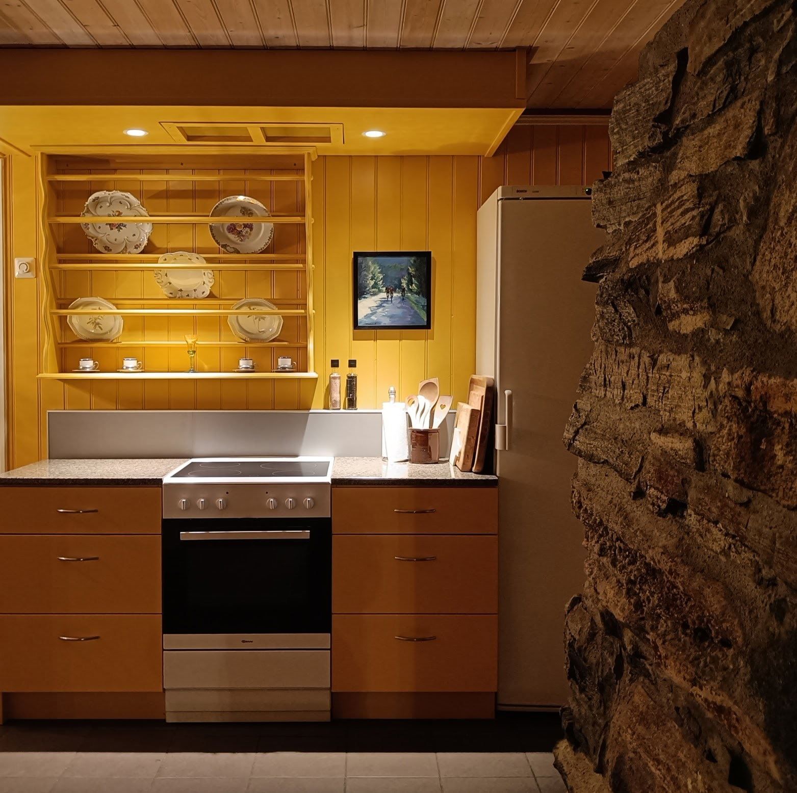 Orheimstunet in Nordfjord - Cozy kitchen with yellow cabinets, open plate shelves, stove and oven, refrigerator, and a rough stone wall..