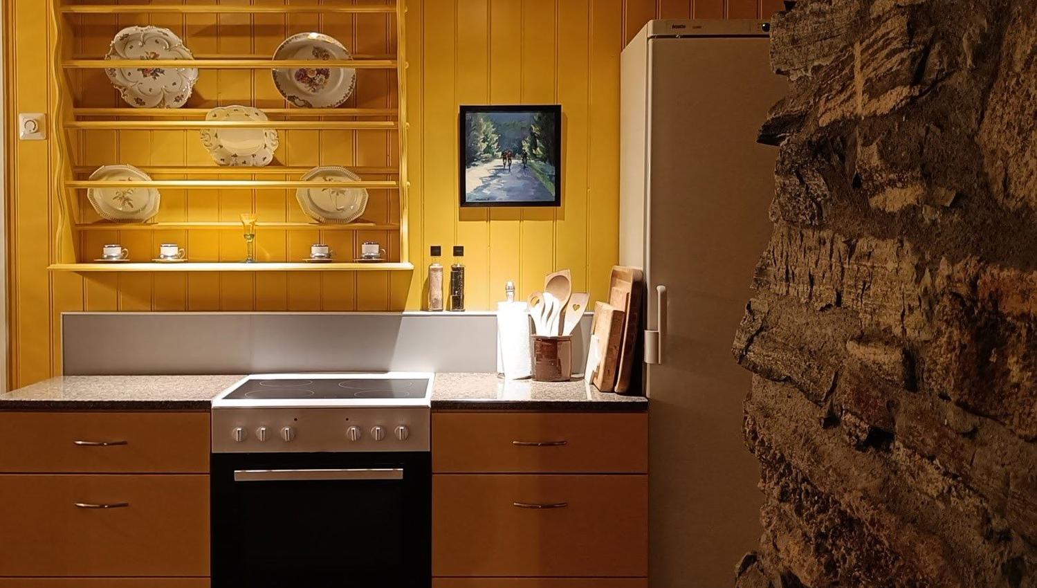 Orheimstunet in Nordfjord - Cozy kitchen with yellow cabinets, open plate shelves, stove and oven, refrigerator, and a rough stone wall..