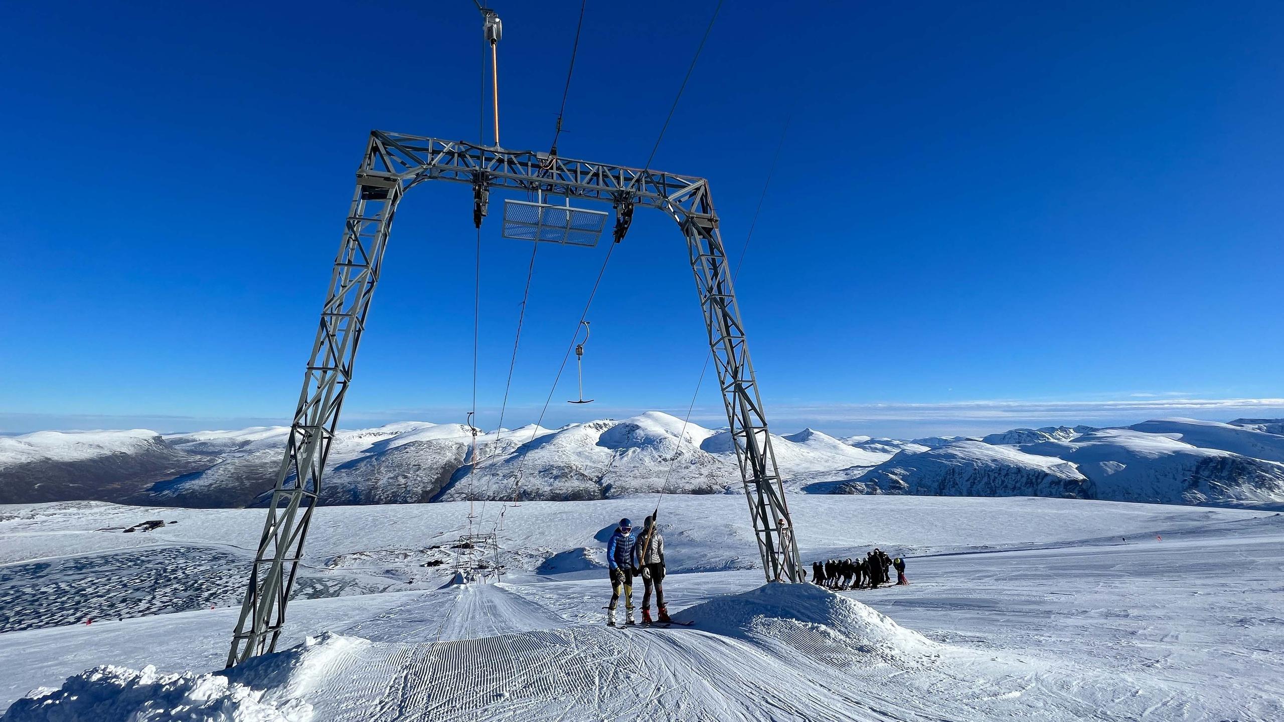 Two skiers stand under a metal ski tow frame on a snowy ridge, with distant mountains beneath a clear blue sky..