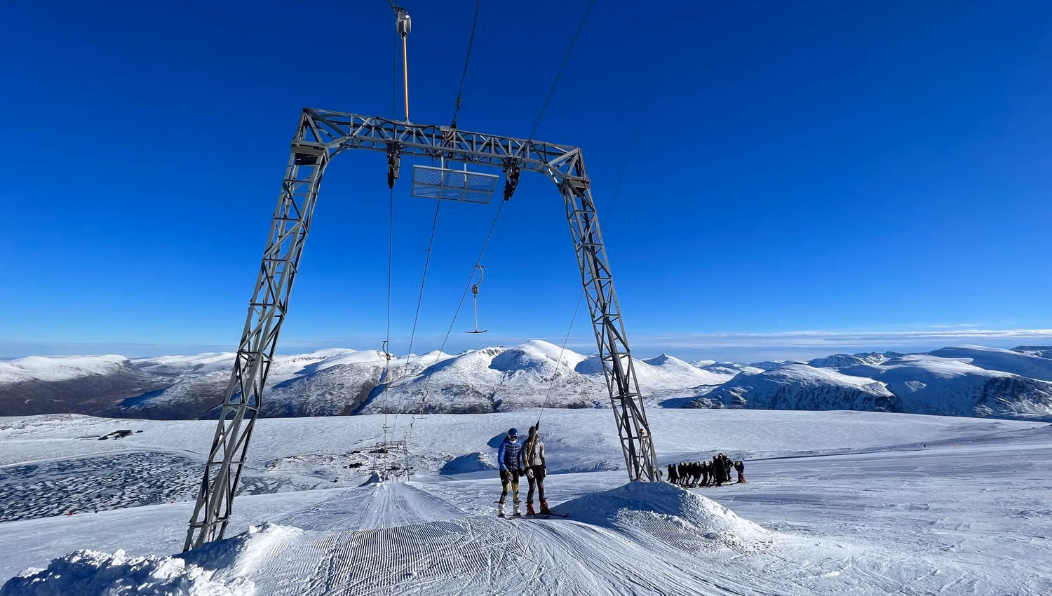 Eit skitrekk med metallportal over ei preparert løype, med to skiløparar og snødekte fjell under blå himmel..
