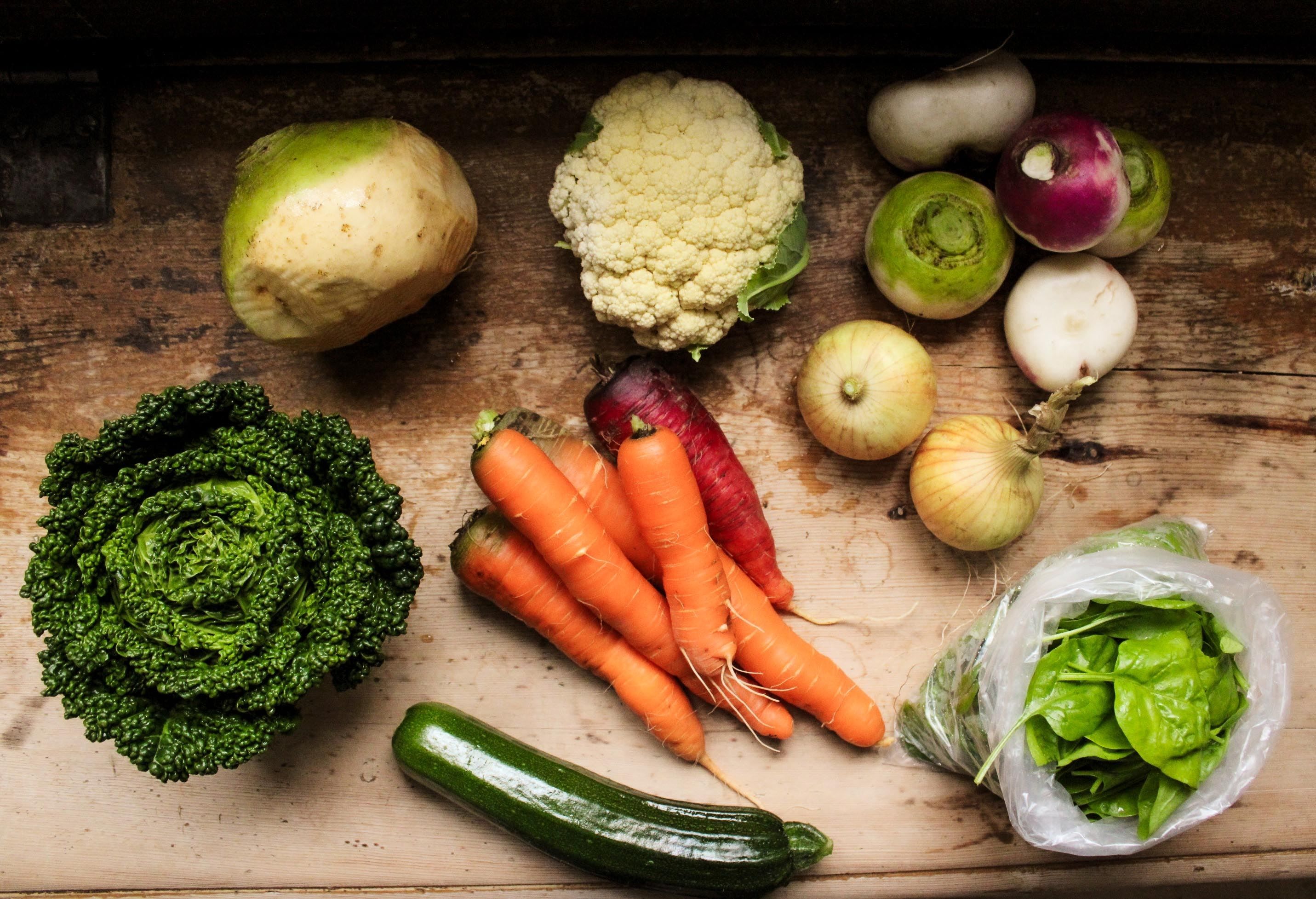 Overhead view of mixed vegetables on a wooden table, including carrots, cauliflower, onions, turnips, kale, zucchini, and spinach..