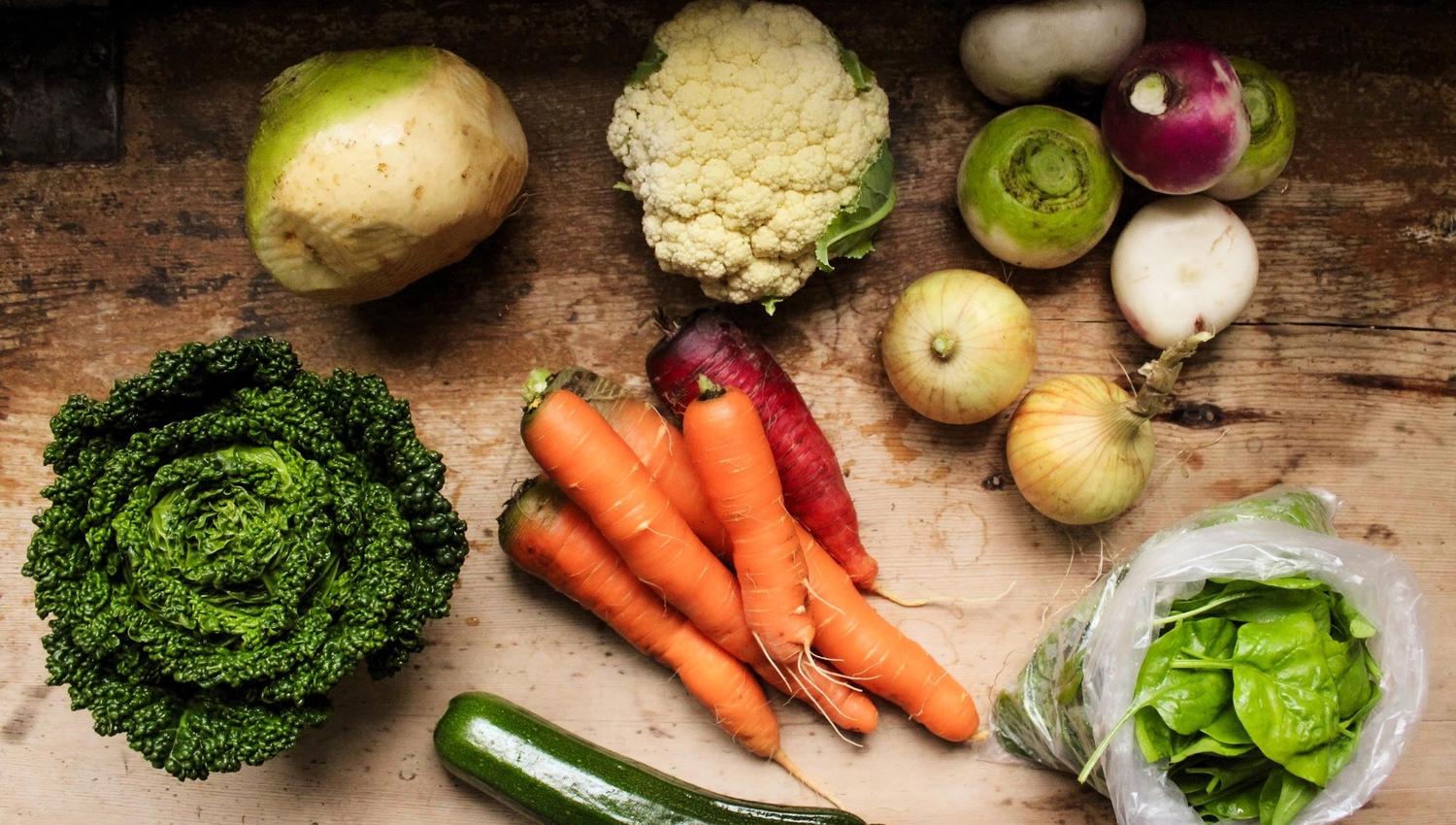 Overhead view of mixed vegetables on a wooden table, including carrots, cauliflower, onions, turnips, kale, zucchini, and spinach..