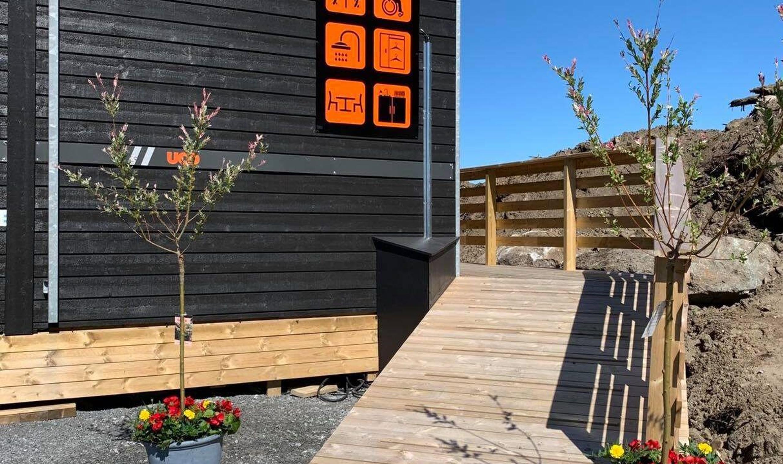 Wooden ramp with railing leads to a dark-sided building with orange icon signage, small trees, and flowers.
