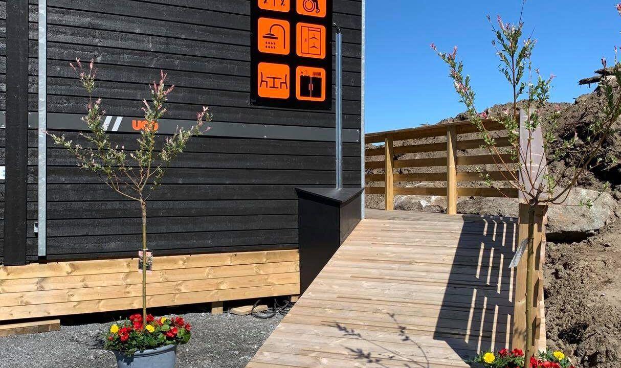 Wooden ramp with railing leads to a dark-sided building with orange icon signage, small trees, and flowers.
