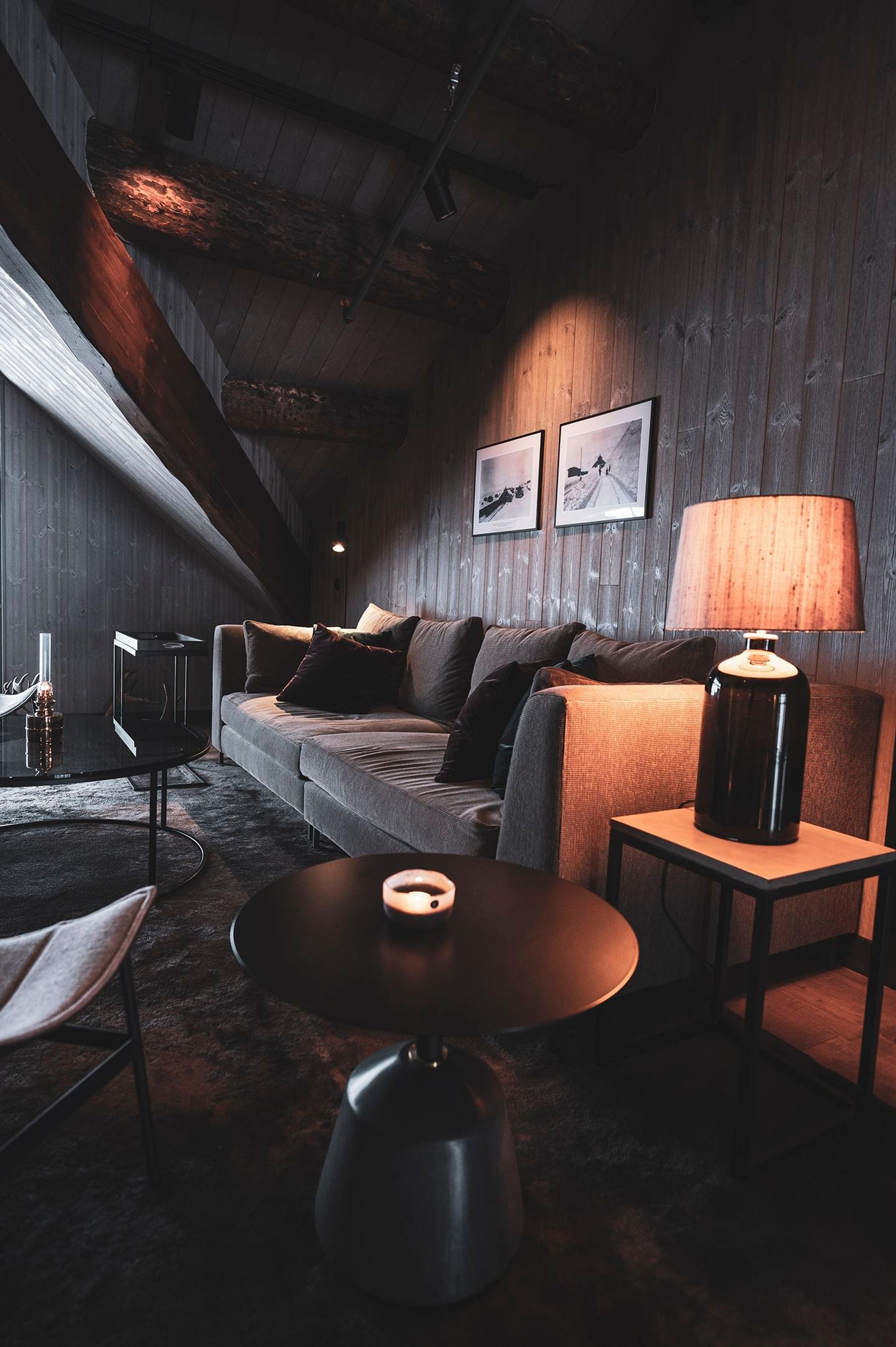 Moody living room with dark wood walls, gray sectional sofa, round tables, framed photos, and a lit table lamp.