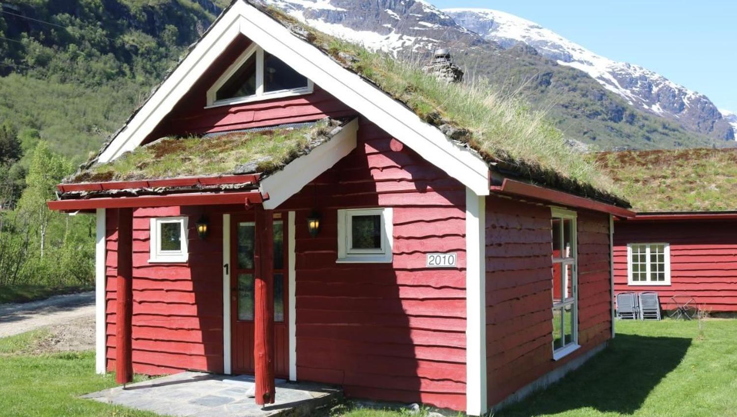Red wooden cabin with white trim and grass-covered roof on a green lawn, with snowy mountains behind, at Aabrekk Gard Trollbu in Oldedalen valley.