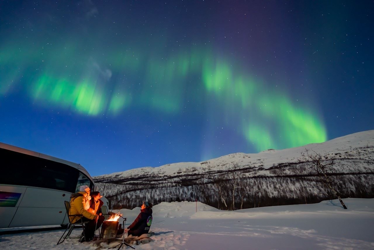 A minibus and guests in a snowy winter landscape beneath the Northern Lights and surrounding mountains.
