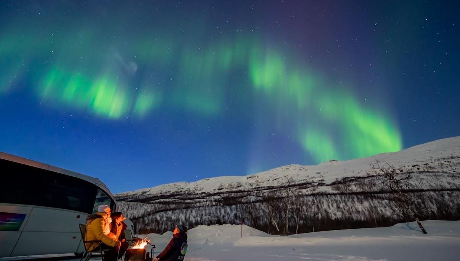 A minibus and guests in a snowy winter landscape beneath the Northern Lights and surrounding mountains.