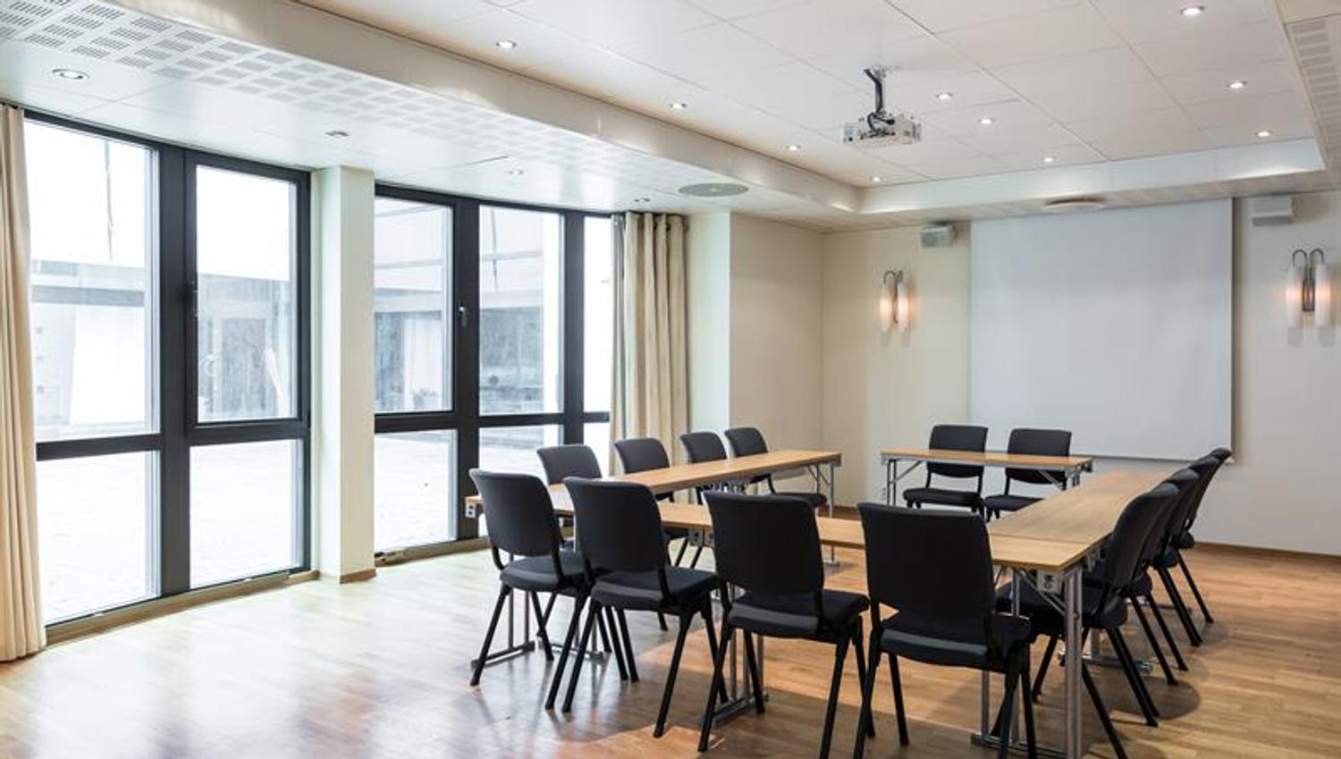 lyst mĂžterom med langbord, stoler og store vinduer som slipper inn dagslys.