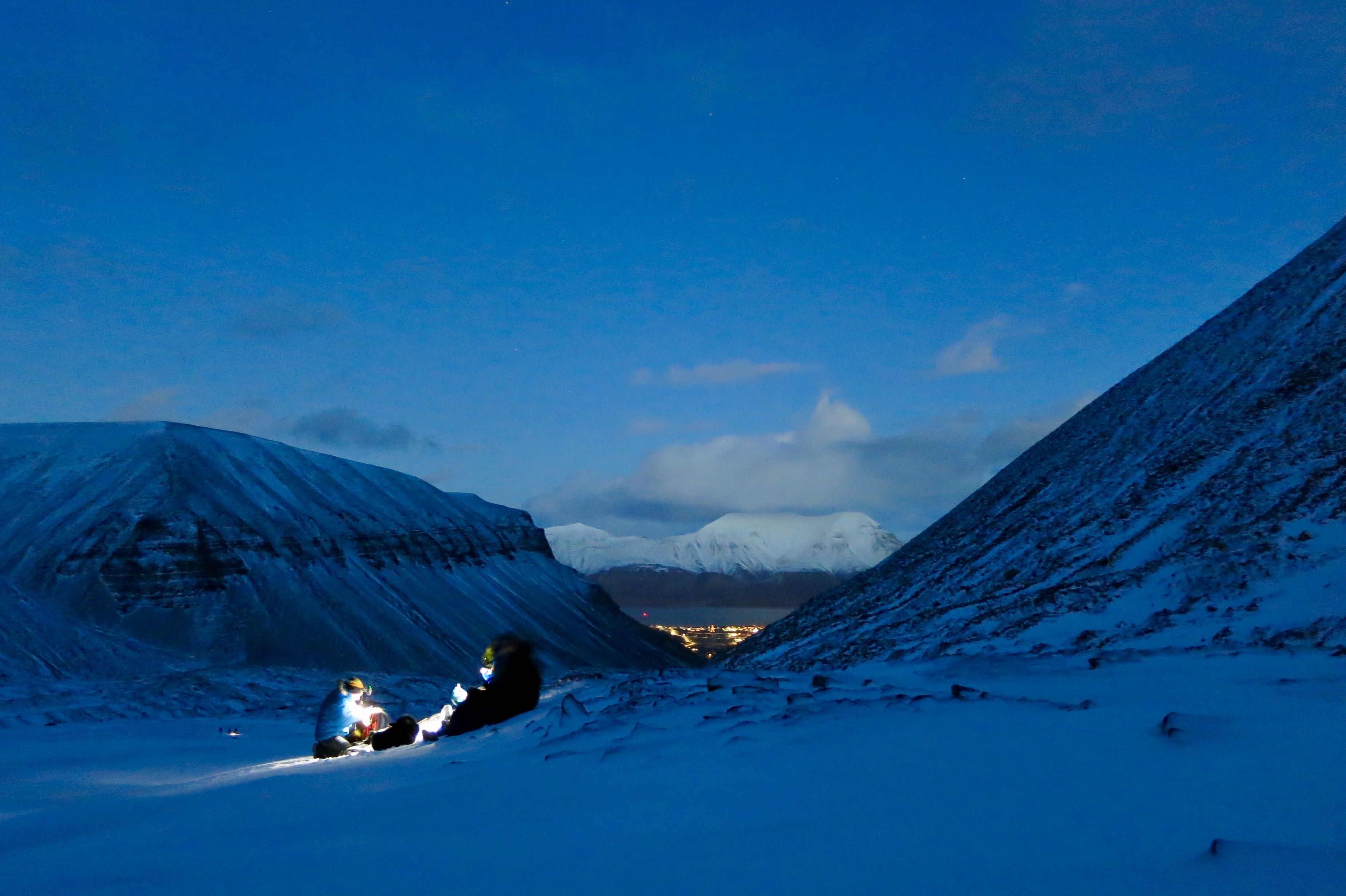 To personer med hodelykt i forgrunnen, med fjell og Longyearbyen i bakgrunnen.