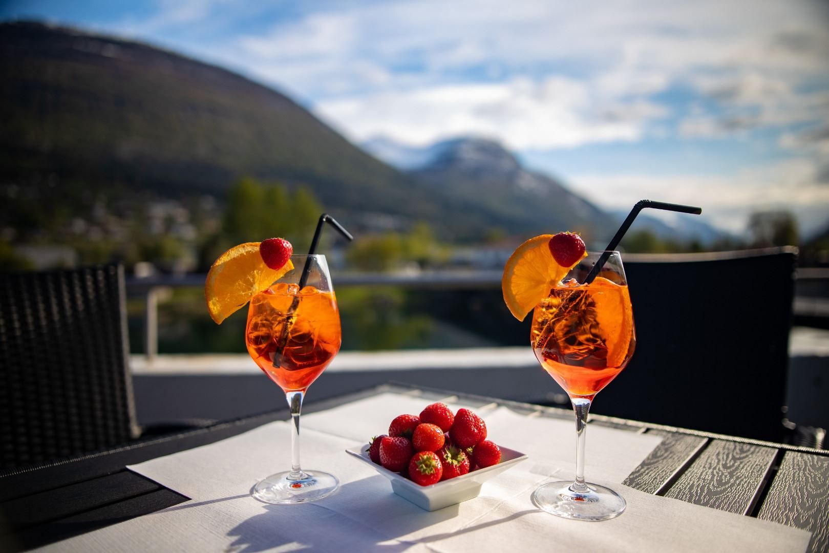 To glas med oransje drink, appelsinskiver og sugerøyr på bord, med jordbærskål og fjellutsikt i bakgrunnen..