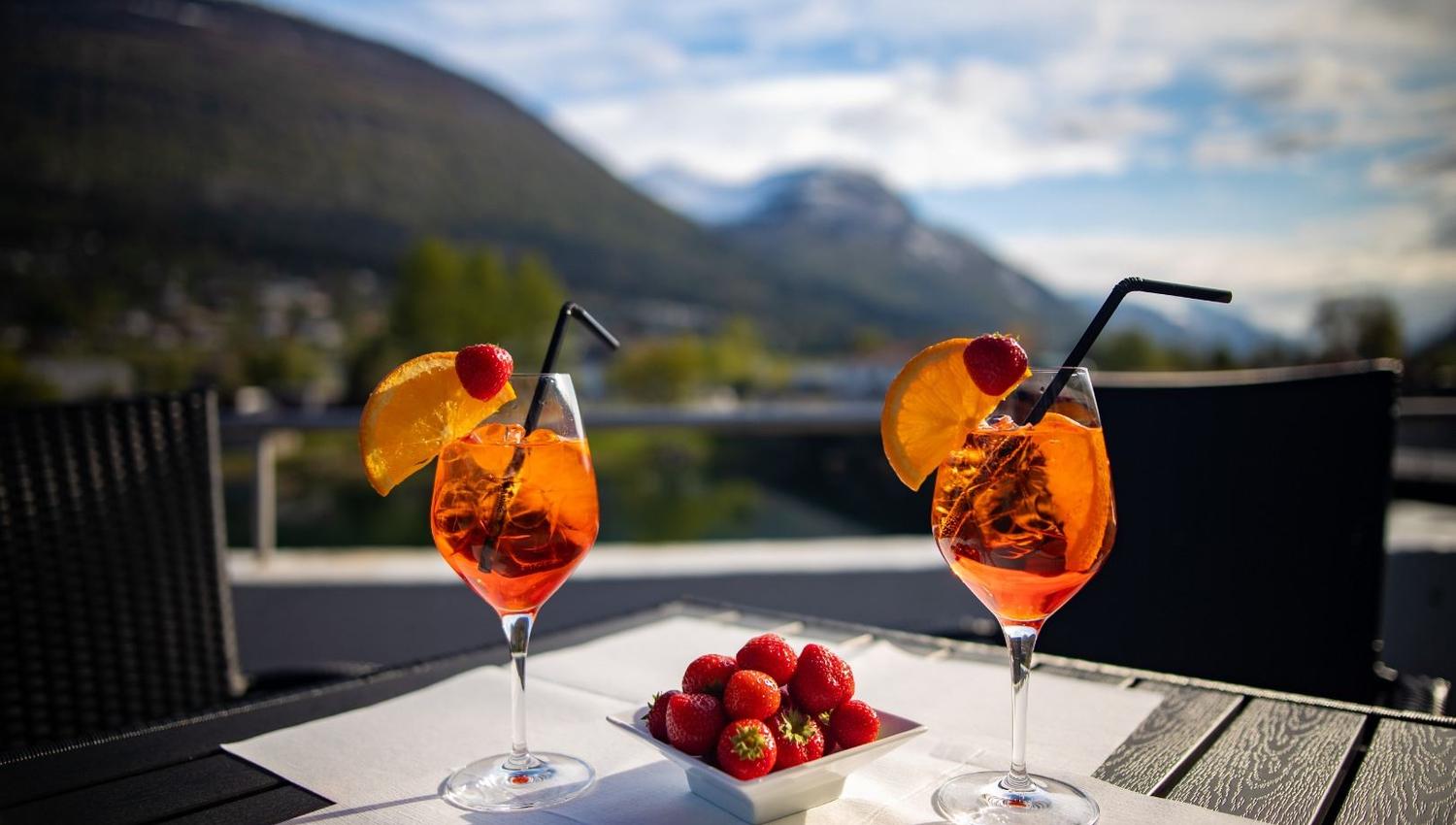 To glas med oransje drink, appelsinskiver og sugerøyr på bord, med jordbærskål og fjellutsikt i bakgrunnen..