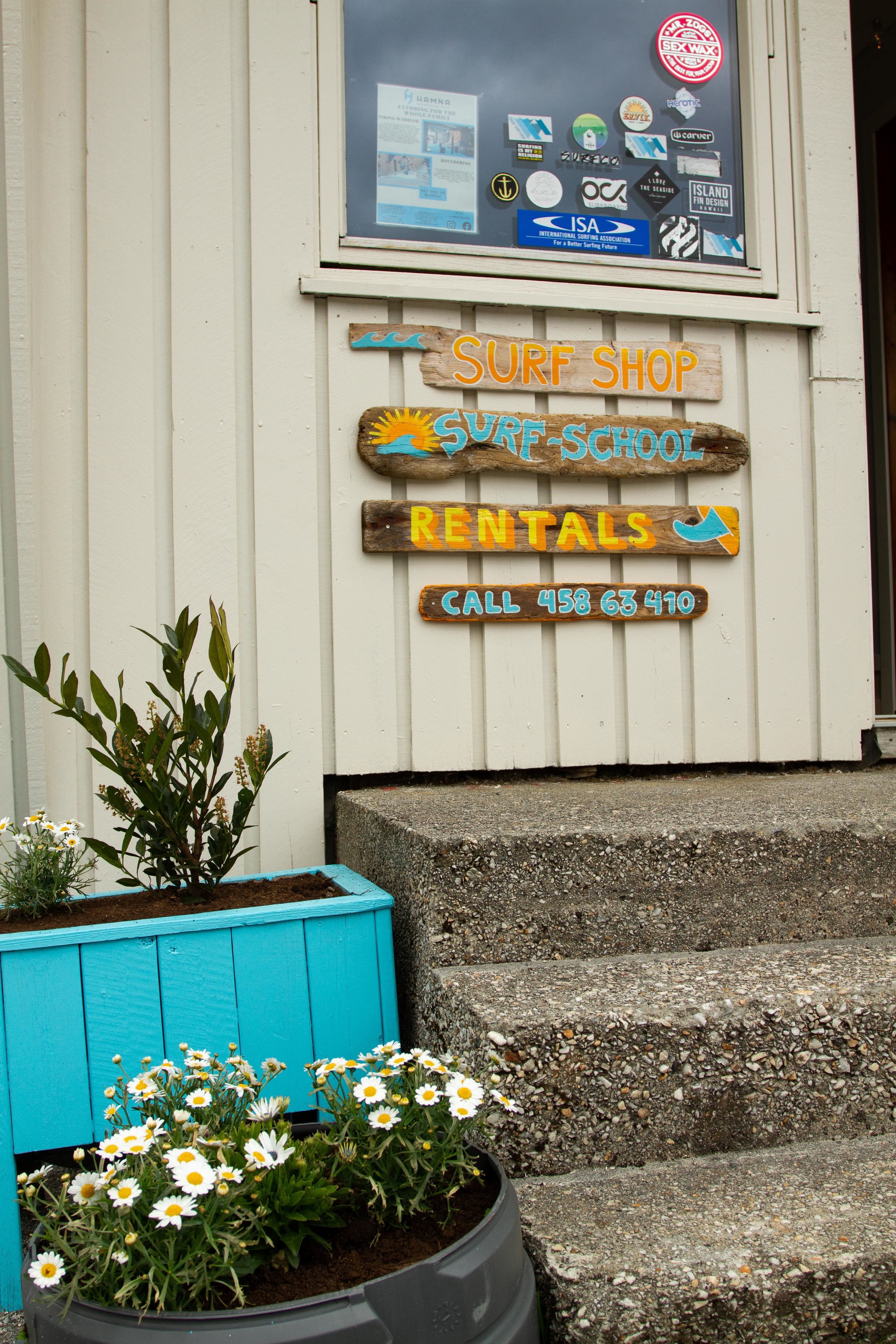 Trepåskilt med «Surf Shop», «Surf-School» og «Rentals» på kvit vegg ved trapp, med blomsterkrukker ved Ervik Surfshop..