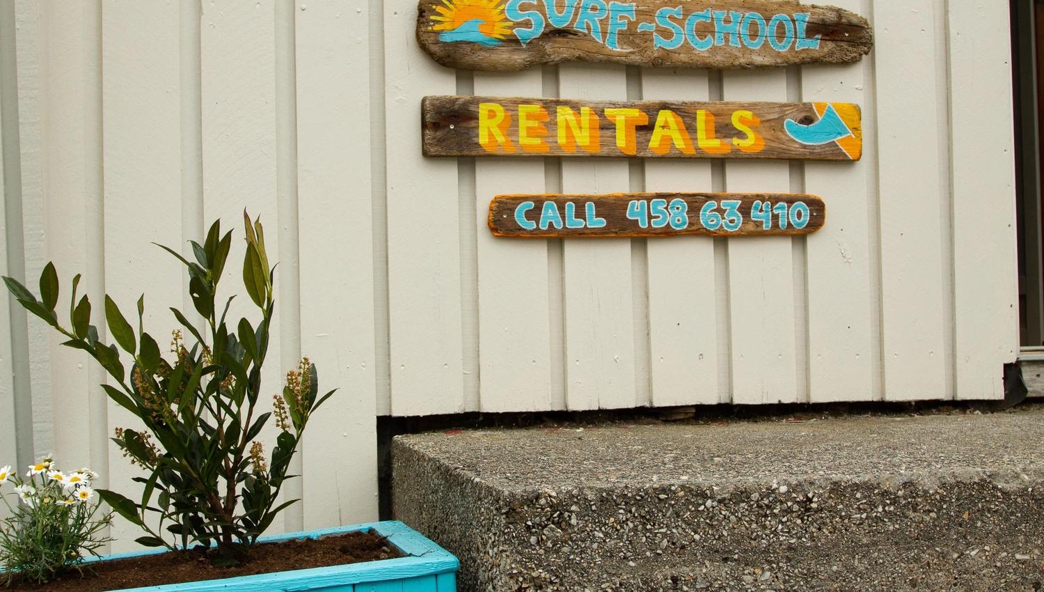 Trepåskilt med «Surf Shop», «Surf-School» og «Rentals» på kvit vegg ved trapp, med blomsterkrukker ved Ervik Surfshop..
