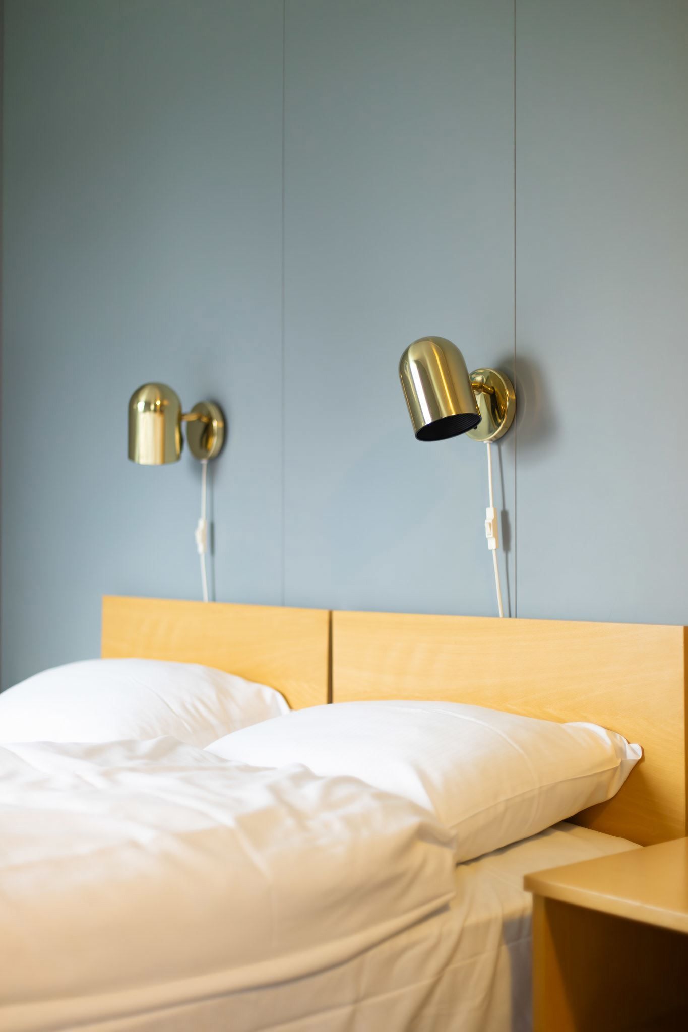 White bed with pillows and wooden headboard beneath two brass wall lamps on a light blue wall in a calm room..
