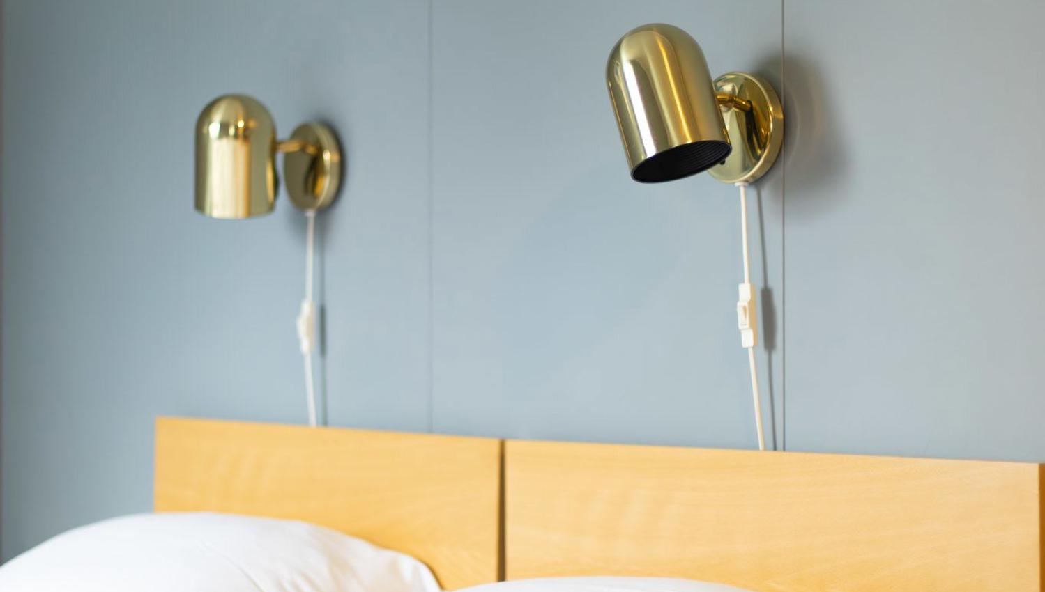 White bed with pillows and wooden headboard beneath two brass wall lamps on a light blue wall in a calm room..
