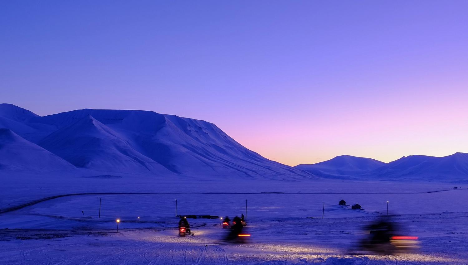 Persons on snowmobiles heading out on a trip with a mountainous landscape covered in blue light in the background