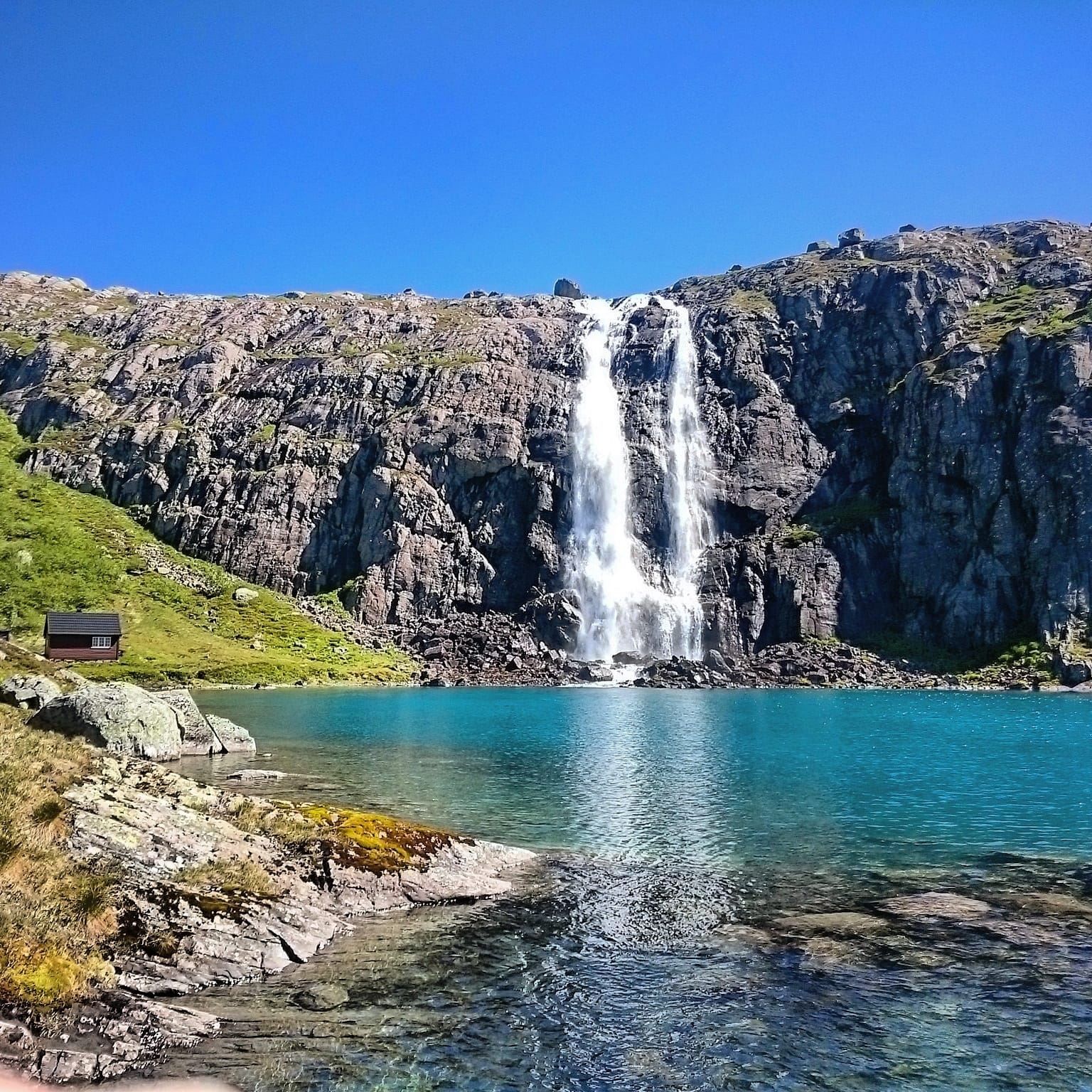 Opplevingar i nærleiken av Bremanger Fjordhotell: Ein høg foss renn ned ei bratt fjellside til eit turkist vatn, med steinstrand og klar blå himmel..