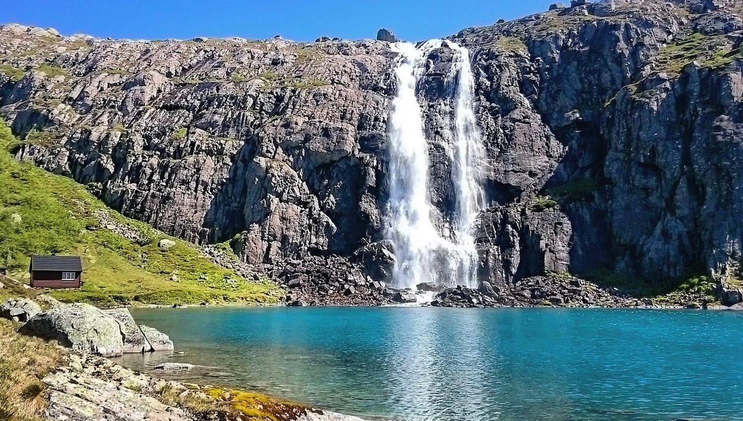 Opplevingar i nærleiken av Bremanger Fjordhotell: Ein høg foss renn ned ei bratt fjellside til eit turkist vatn, med steinstrand og klar blå himmel..