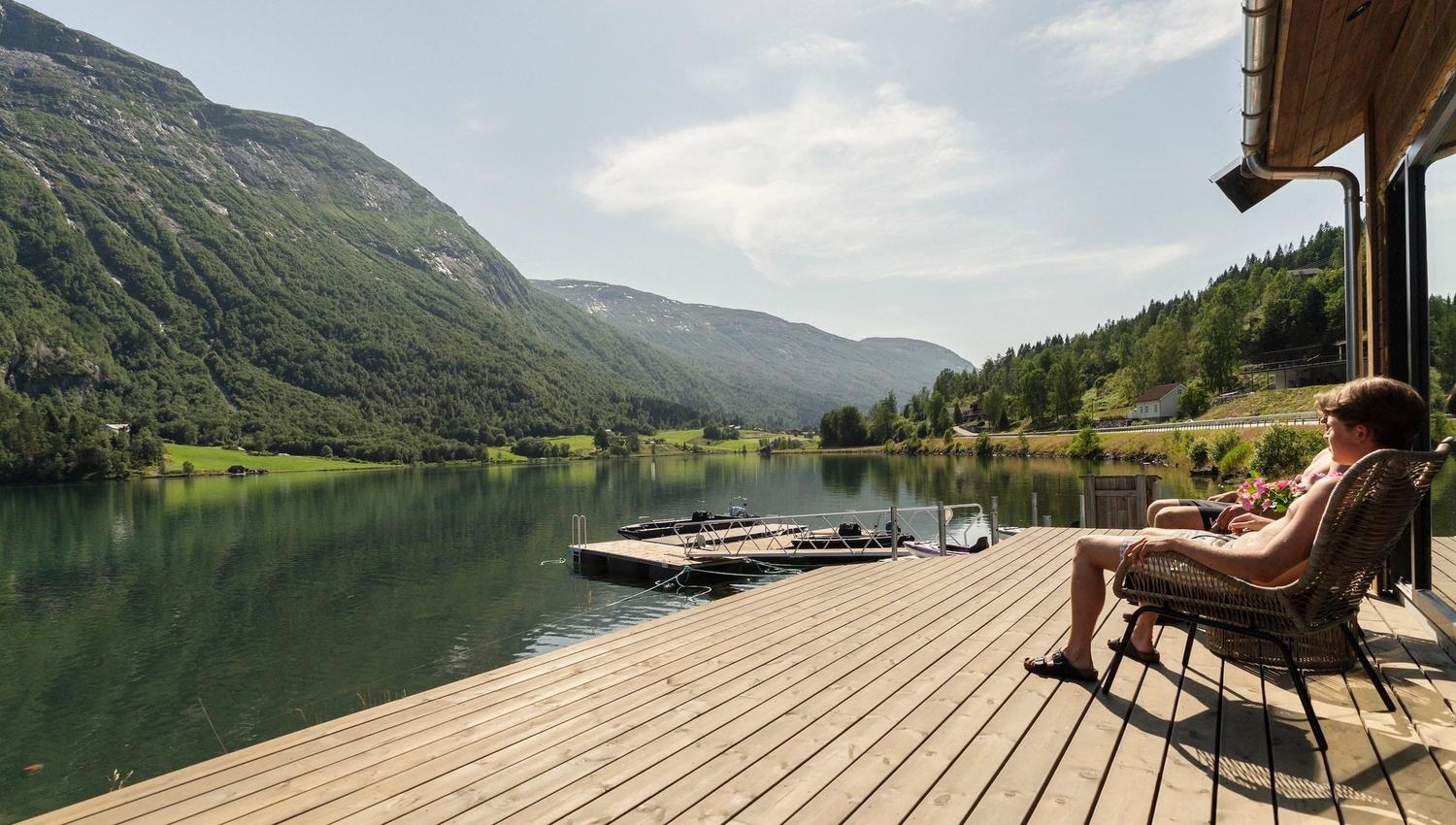 Two people sit in wicker chairs on a wooden deck overlooking a calm lake, docked boats, and green mountains..