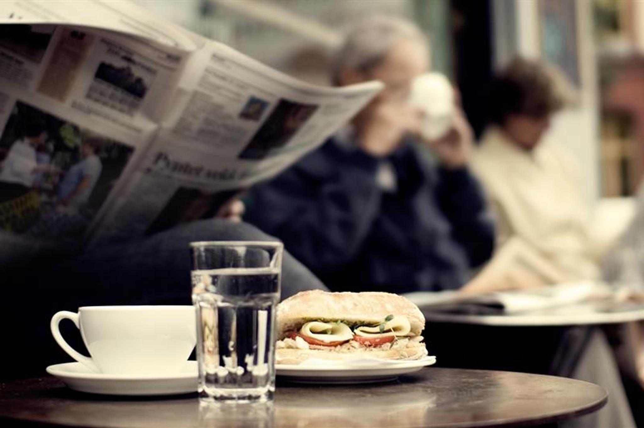 Detaljbilde av en kaffekopp, et glass vann og en ciabatta med person som leser en avis i bakgrunnen.