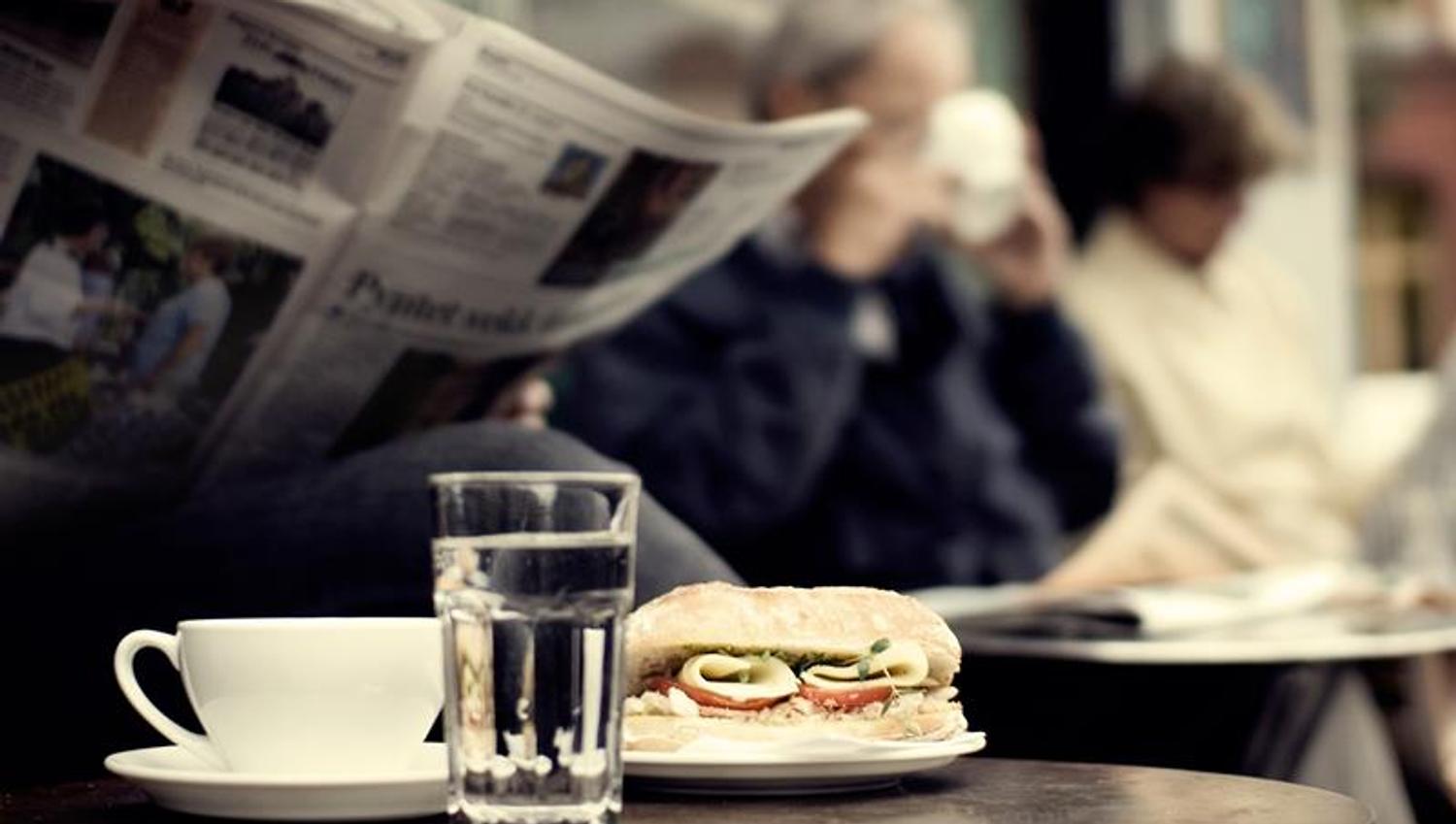 Detaljbilde av en kaffekopp, et glass vann og en ciabatta med person som leser en avis i bakgrunnen.