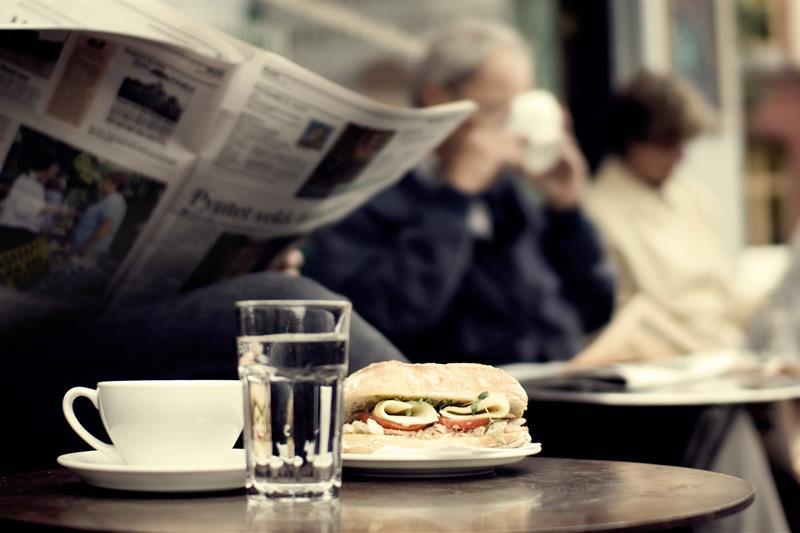 Detaljbilde av en kaffekopp, et glass vann og en ciabatta med person som leser en avis i bakgrunnen.