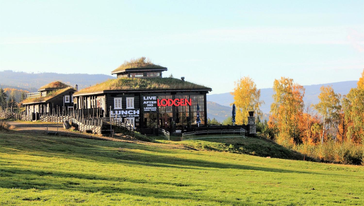 Moderne lodge med torvtak på en gresskledd ås, omgitt av høsttrær, med fjell i bakgrunnen..